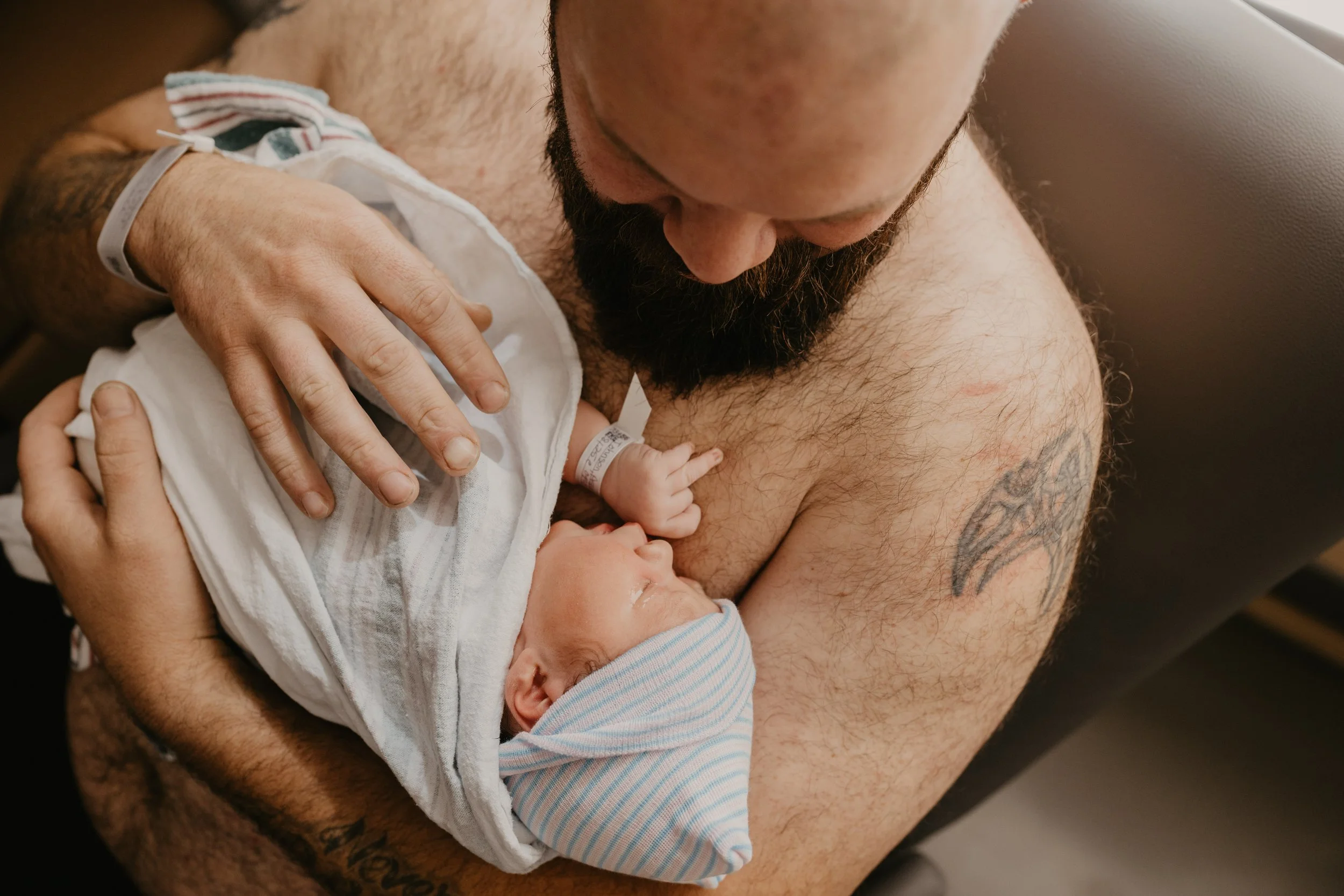 A man with a beard and tattoos holding a newborn baby wrapped in a hospital blanket on his chest, looking down at the baby.