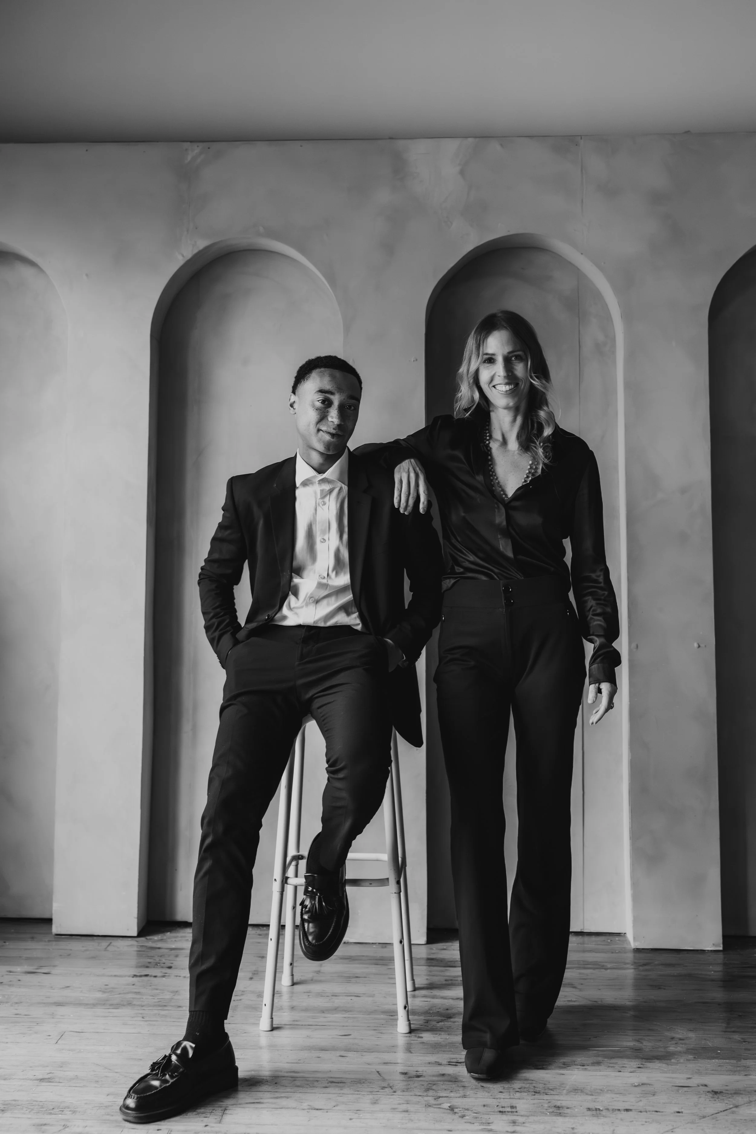 Black and white photo of a young man and woman dressed in formal attire, posing together indoors. The man is sitting on a stool, wearing a suit and tie, with one hand in his pocket. The woman is standing beside him, smiling, with her arm resting on h