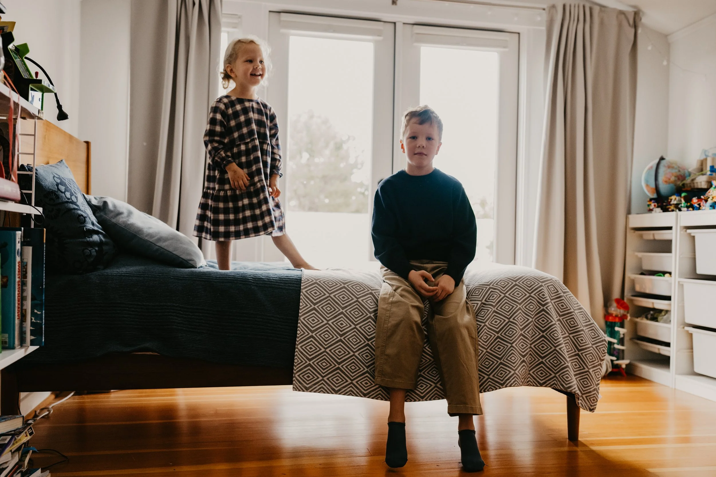 Two children, a girl and a boy, inside a bedroom with large windows in the background. The girl is standing on the bed, smiling, wearing a checked dress. The boy is sitting on the edge of the bed, looking at the camera, wearing a dark sweater and bei