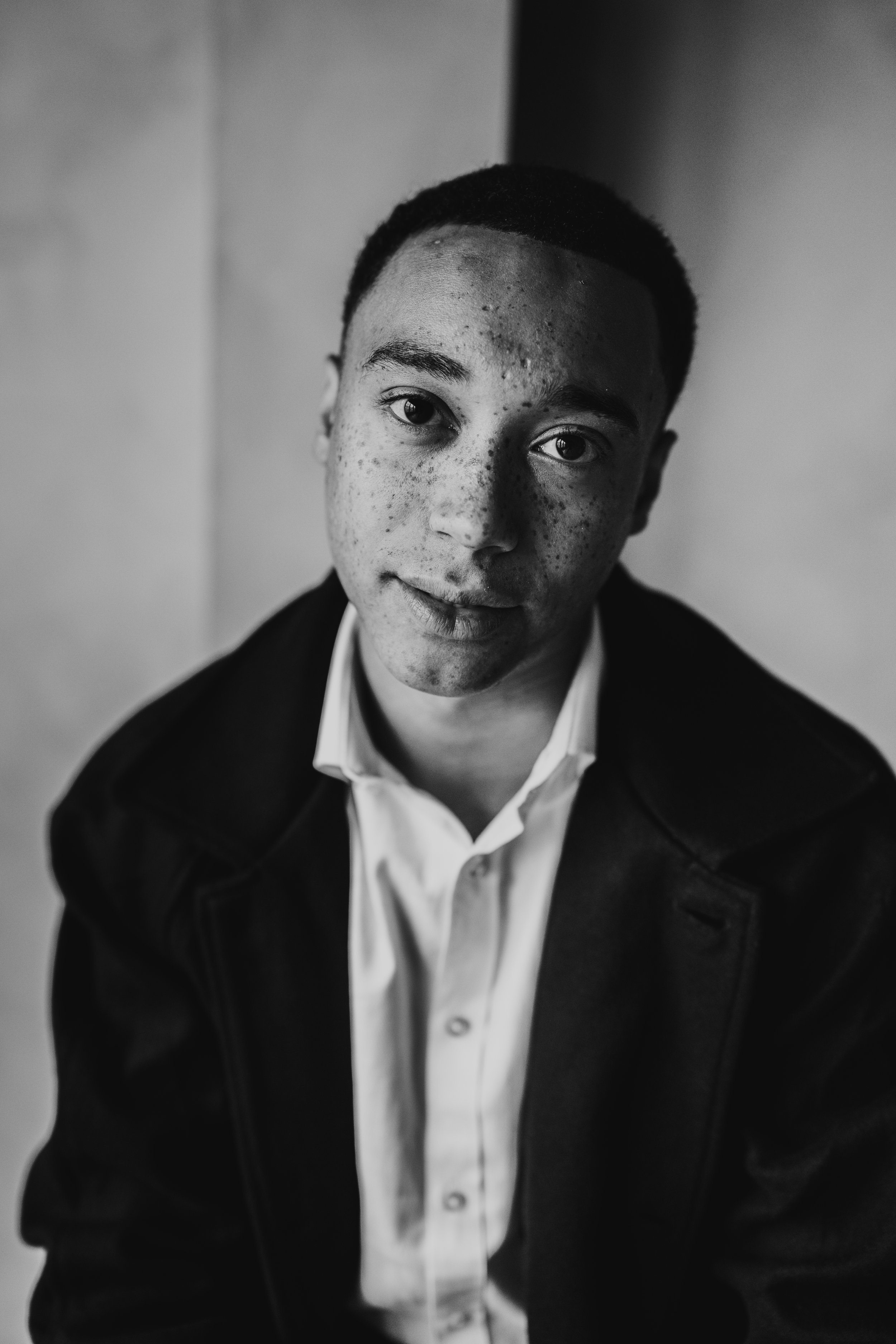 A black-and-white portrait of a young man with short hair, wearing a dark jacket and a white shirt, looking directly at the camera.