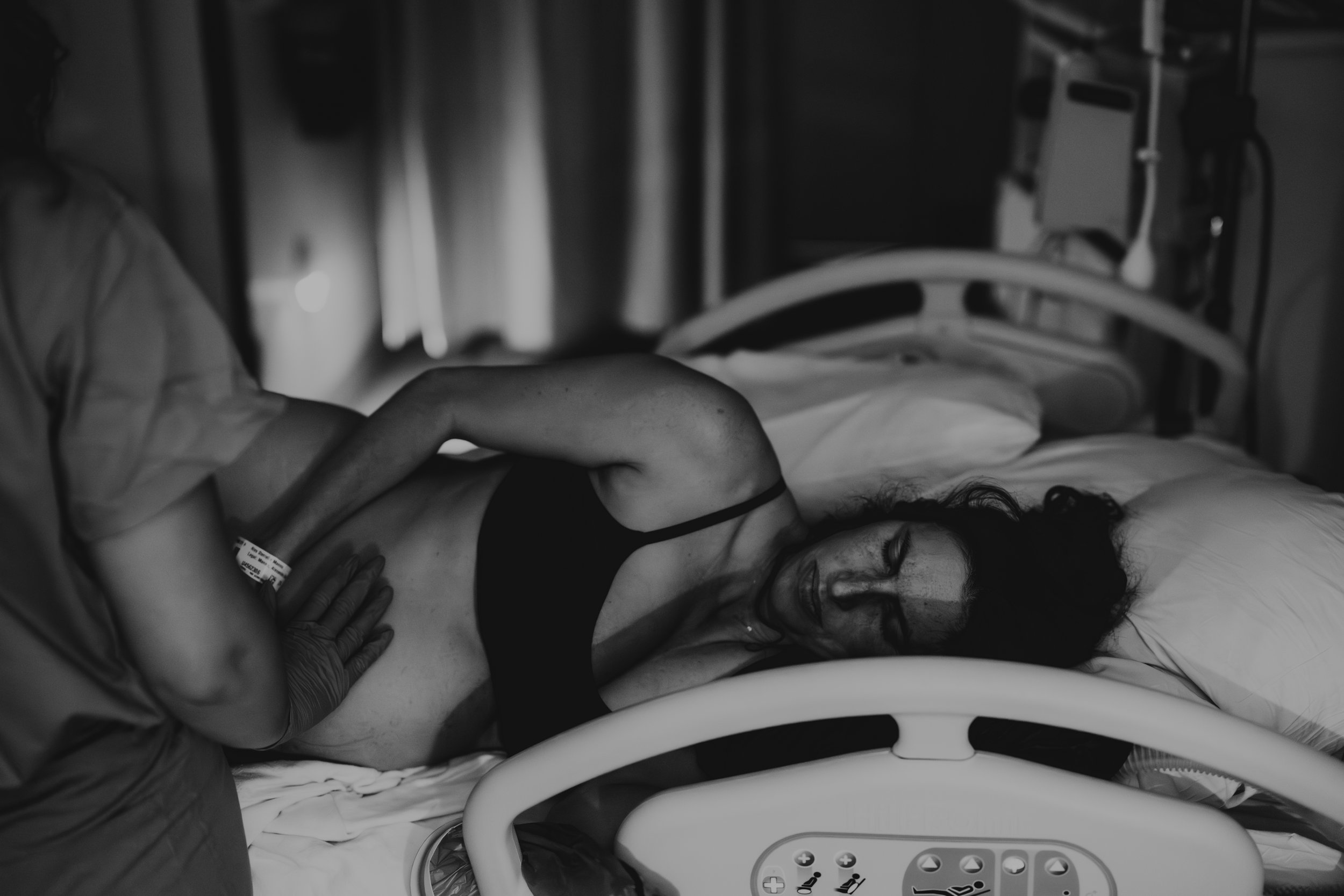 Black and white photo of a woman lying in a hospital bed, visibly distressed, with a healthcare worker’s hand on her abdomen.