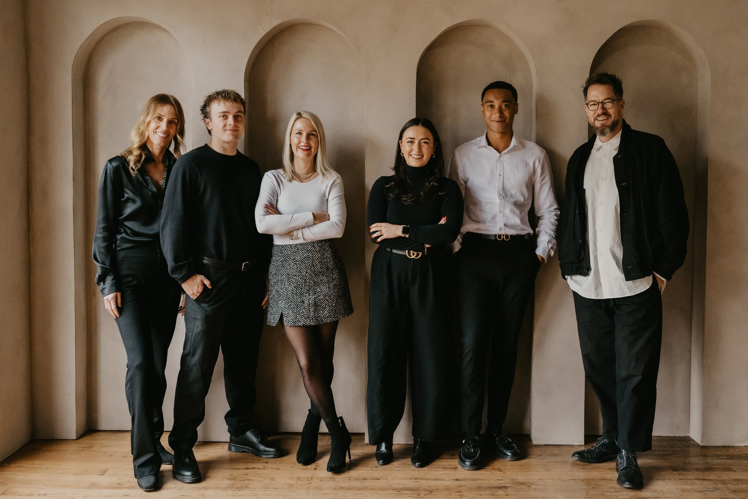 Group of seven diverse professionals standing indoors against a beige wall with arches, smiling at the camera, dressed in business casual attire.