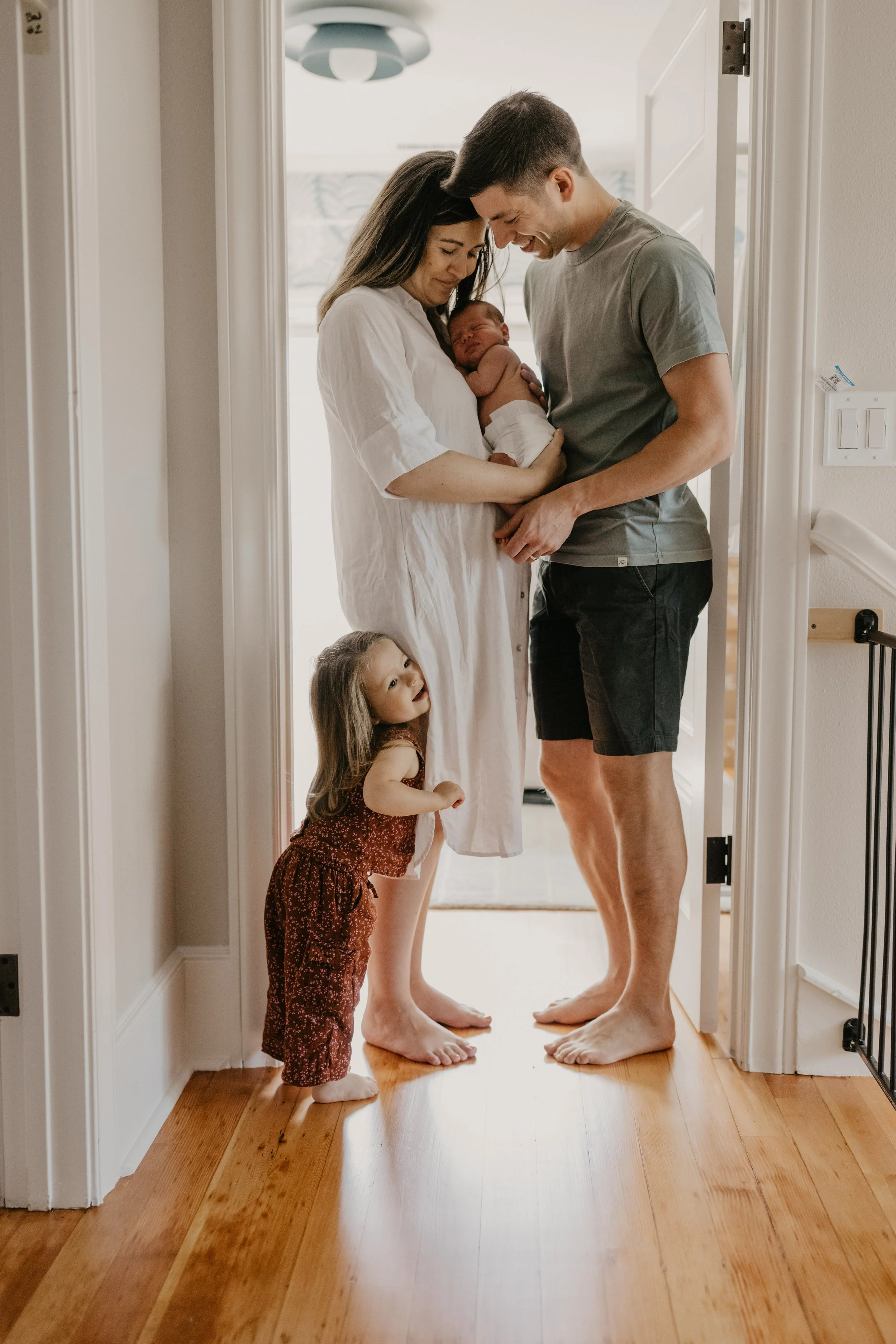 A family standing in a doorway, with a woman holding a newborn baby, a man leaning in close, and a young girl looking up at them. The scene is bright and warm, capturing a tender moment.