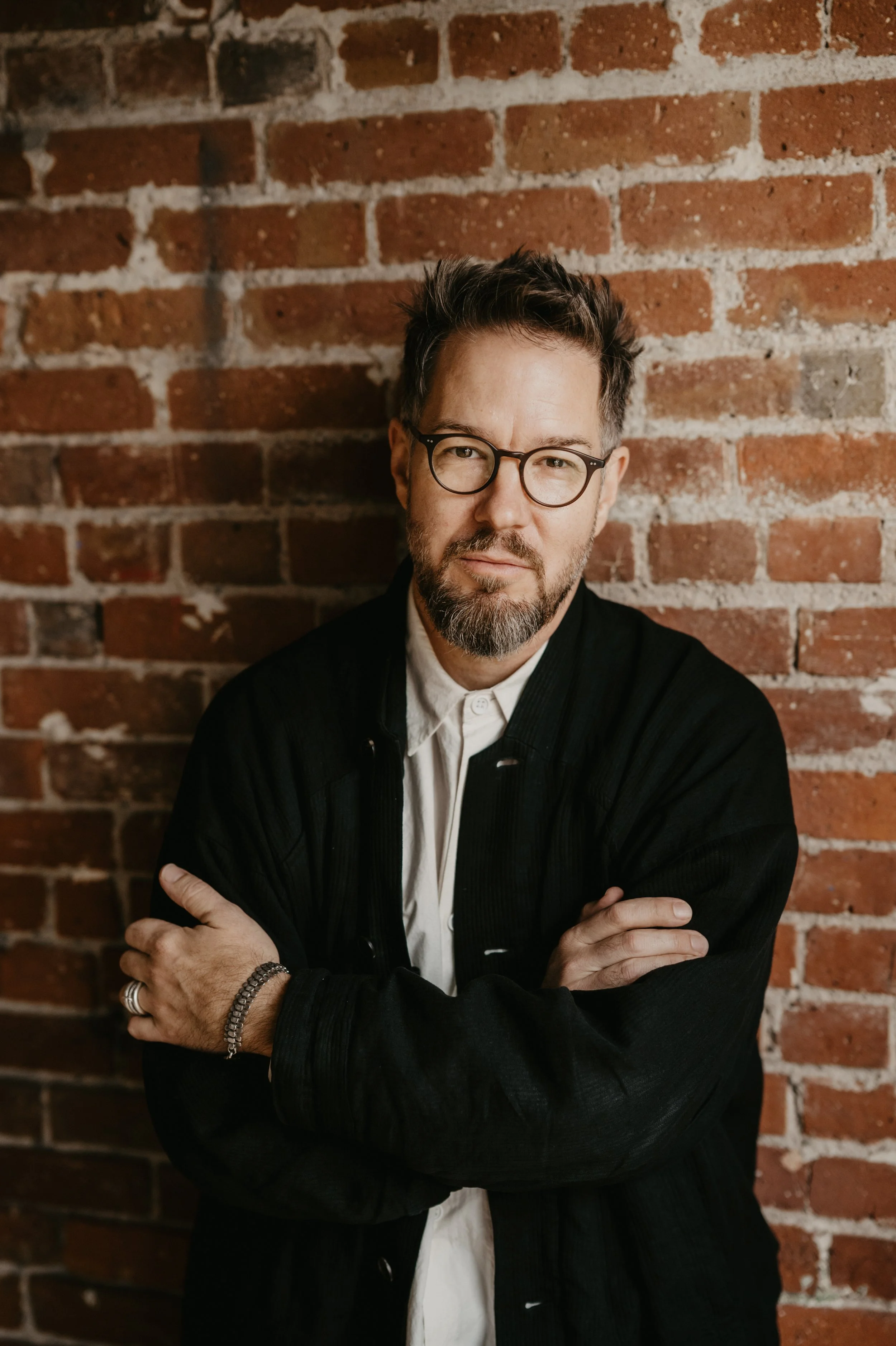 A man with glasses, a beard, and short dark hair stands with arms crossed against a red brick wall. He wears a white shirt and a black jacket.