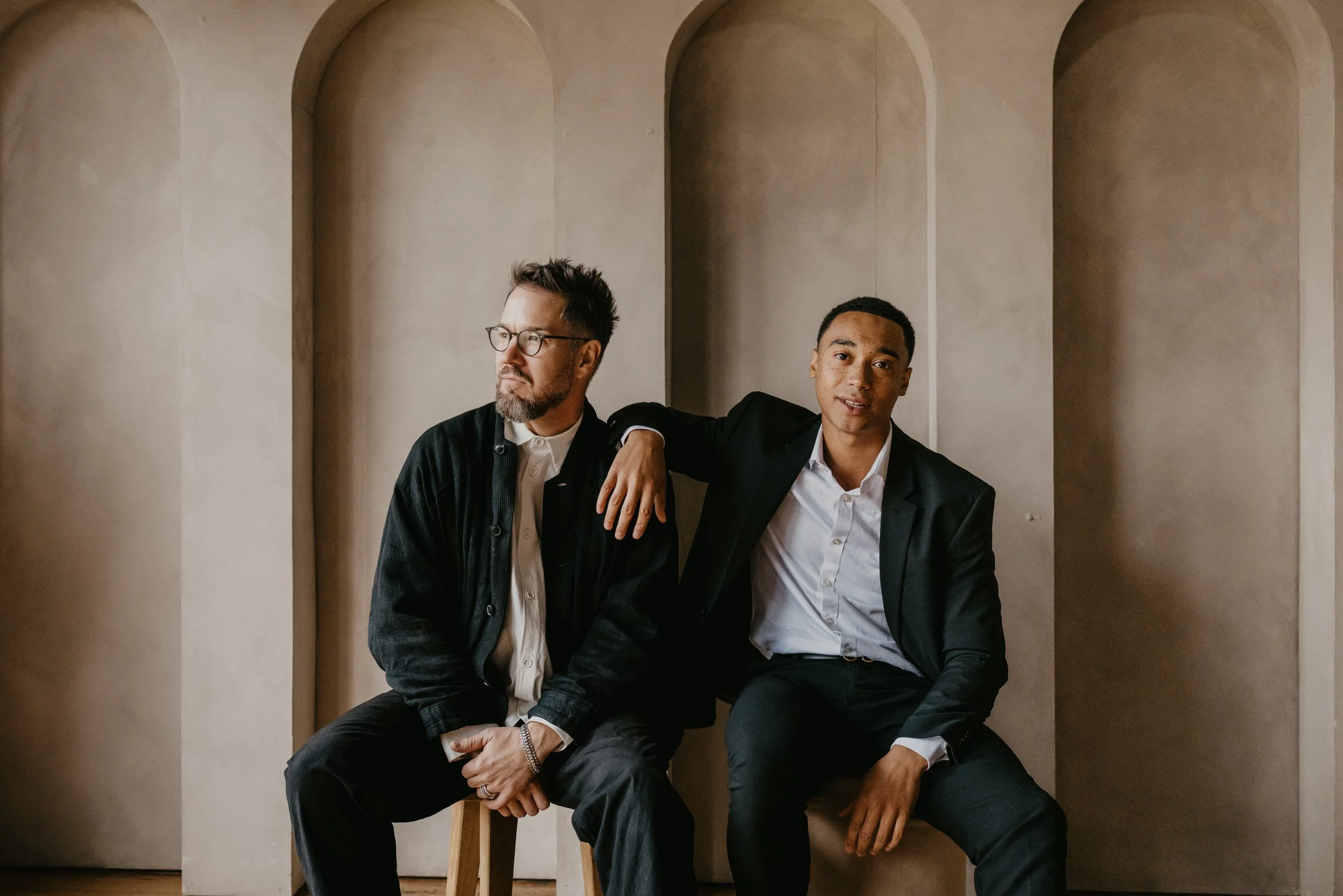Two men sitting on chairs against a neutral wall, one wearing glasses and a casual jacket, the other in a black suit with an open collar. They are in a relaxed pose, with the man in the suit resting his arm on the other's shoulder.