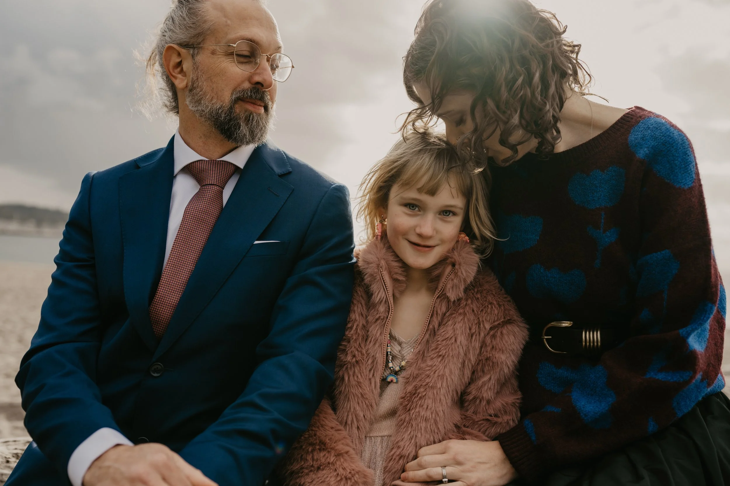A family of three, a man, a woman, and a young girl, sitting outdoors near a beach. The man has gray hair and a beard, wearing glasses and a blue suit with a red tie. The girl has light brown hair, wearing a pink faux fur coat and jewelry. The woman 