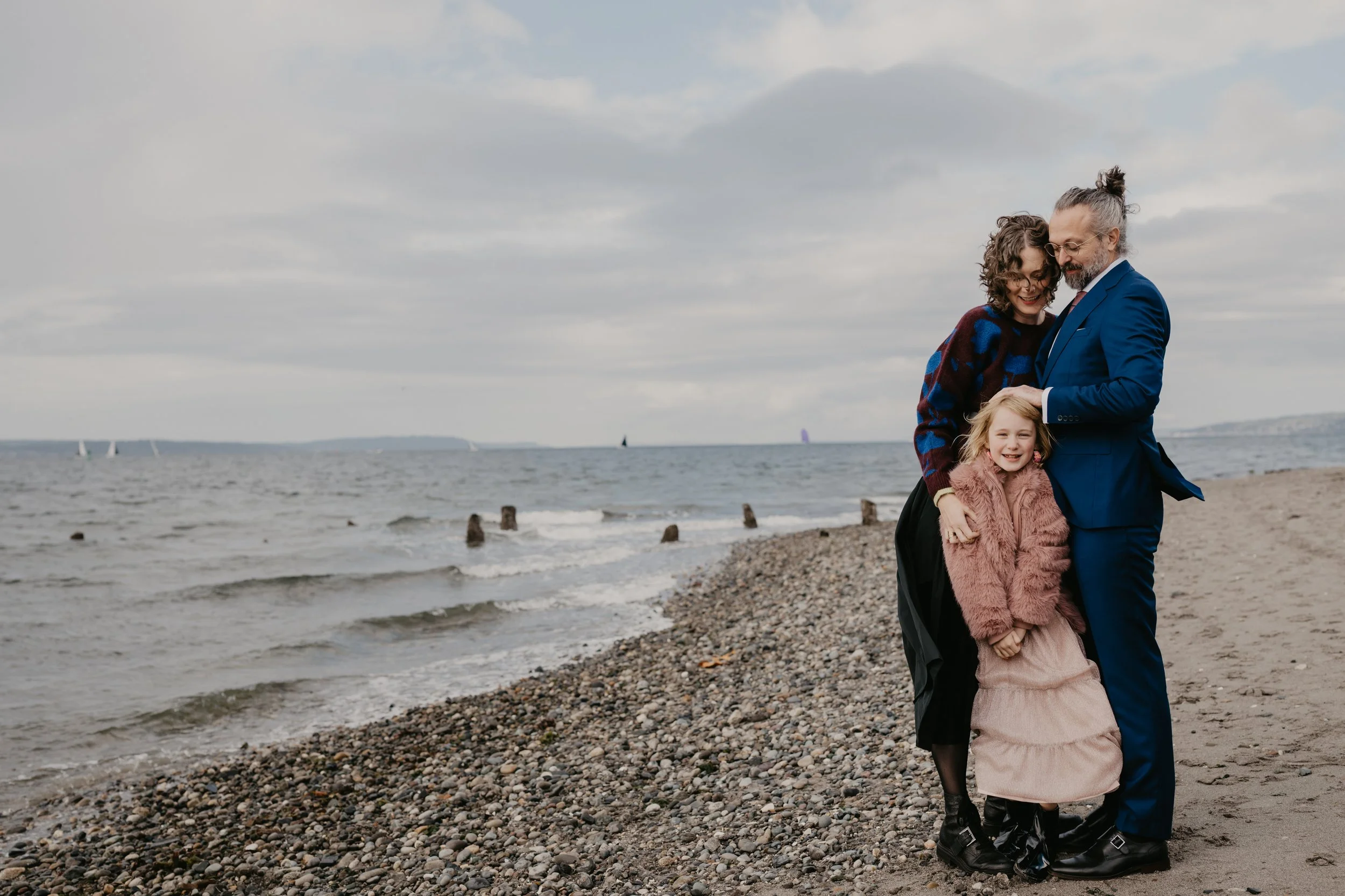 A family of three stands on a pebble beach by the ocean, smiling and hugging, with sailboats in the water and cloudy sky above.