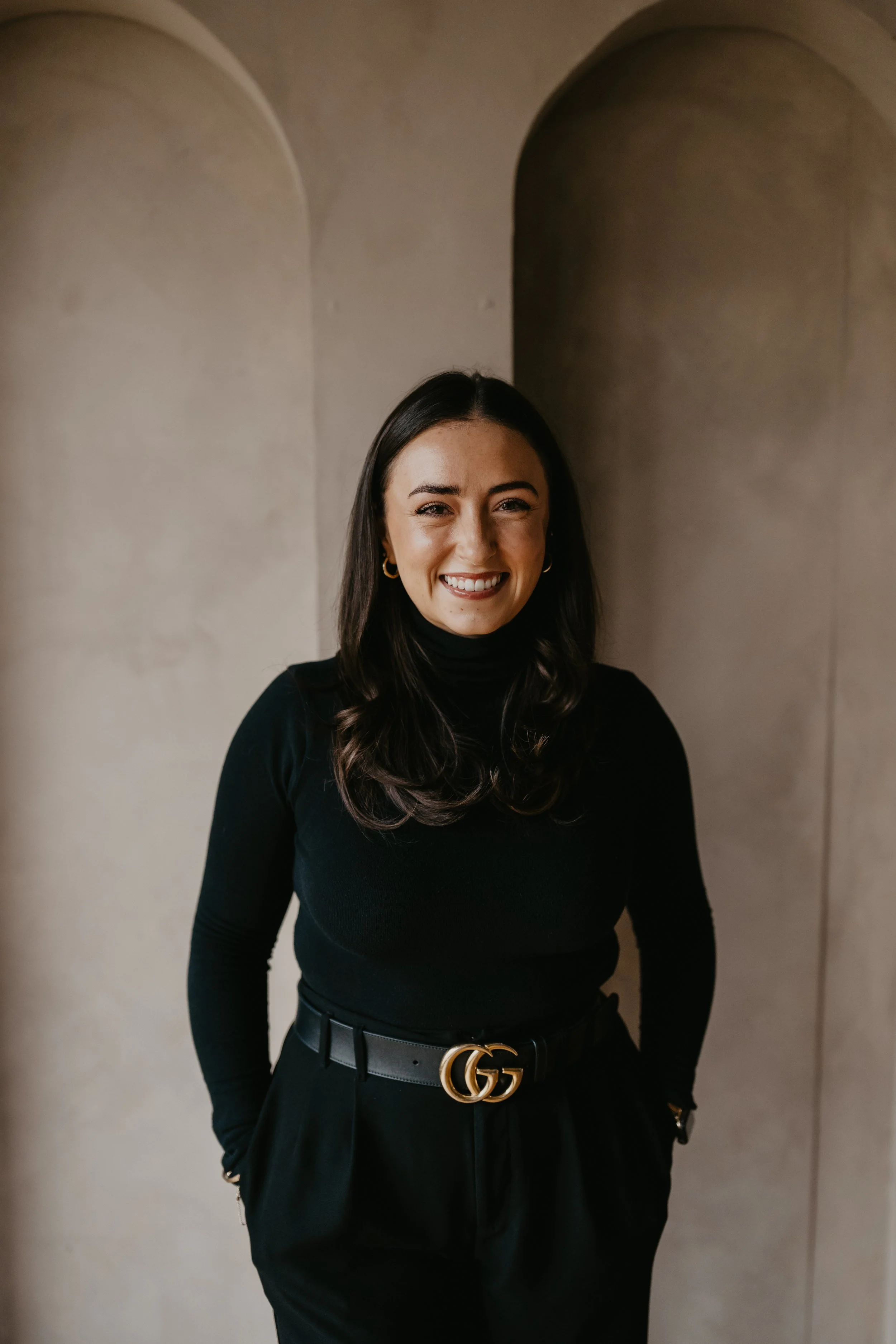 A woman with long dark hair, wearing a black turtleneck and black pants with a Gucci belt, smiling in front of a beige wall.