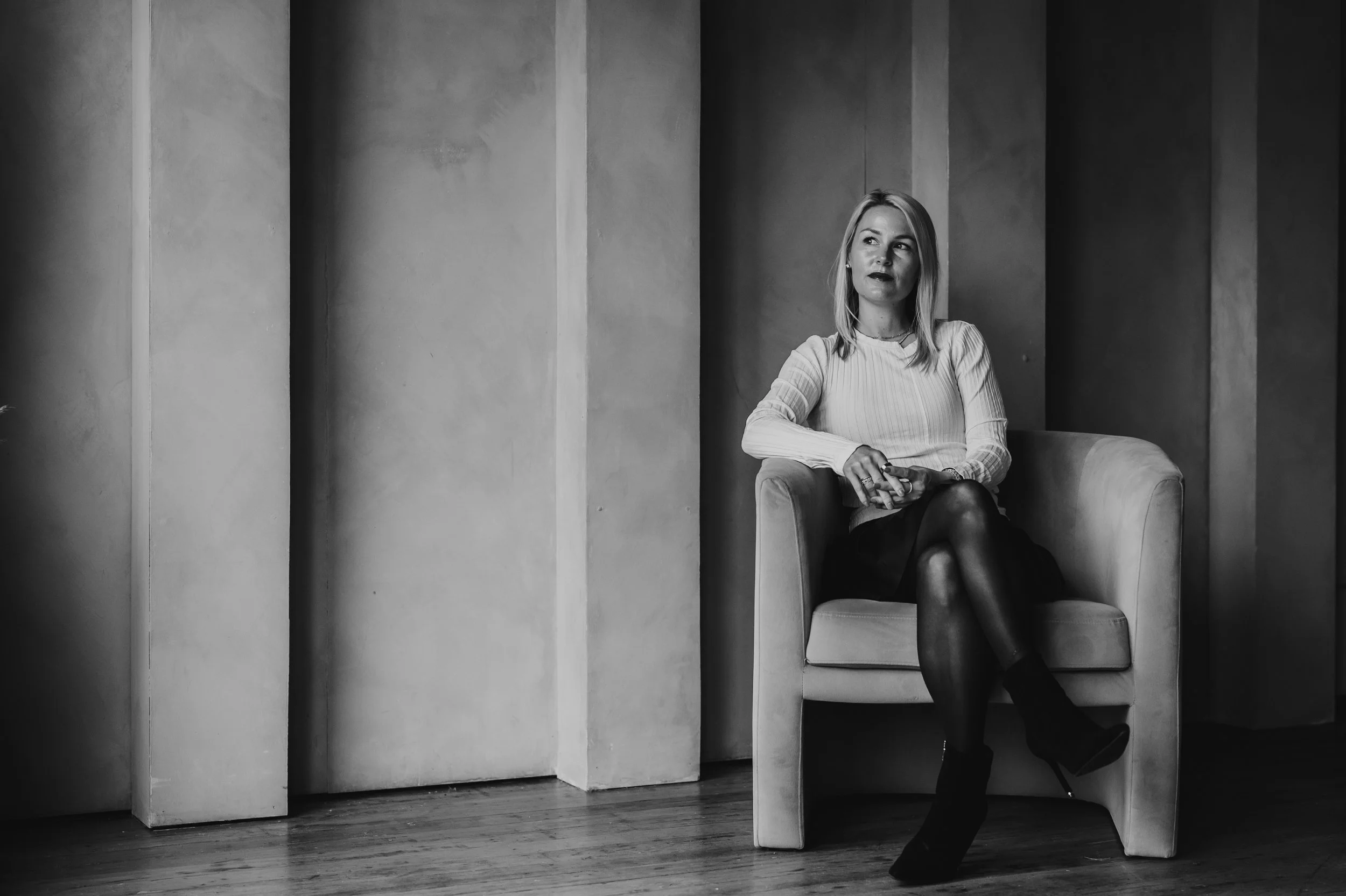 A woman sitting on a sofa in an indoor space, dressed in a white top, black skirt, and black heels, looking to her left in a contemplative pose.