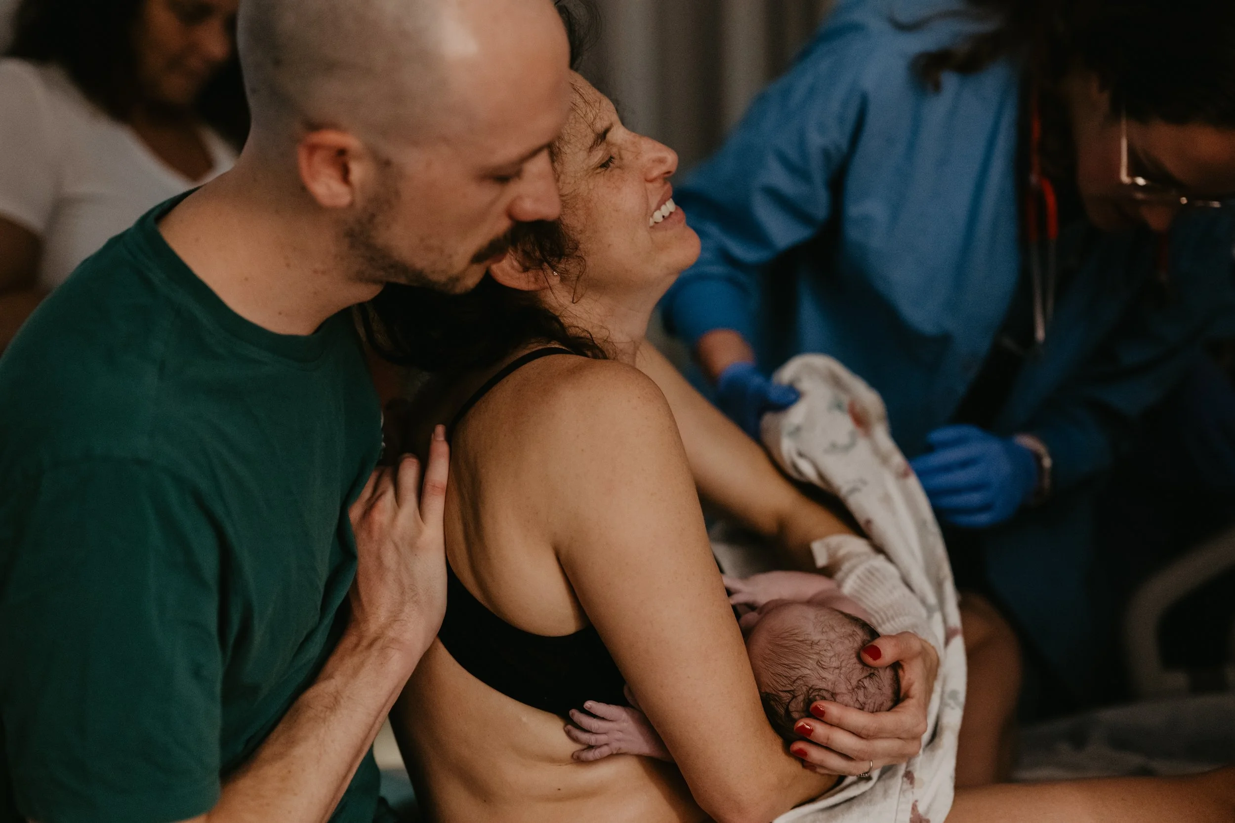 A woman holding a newborn baby while a man leans in close during childbirth in a hospital setting.