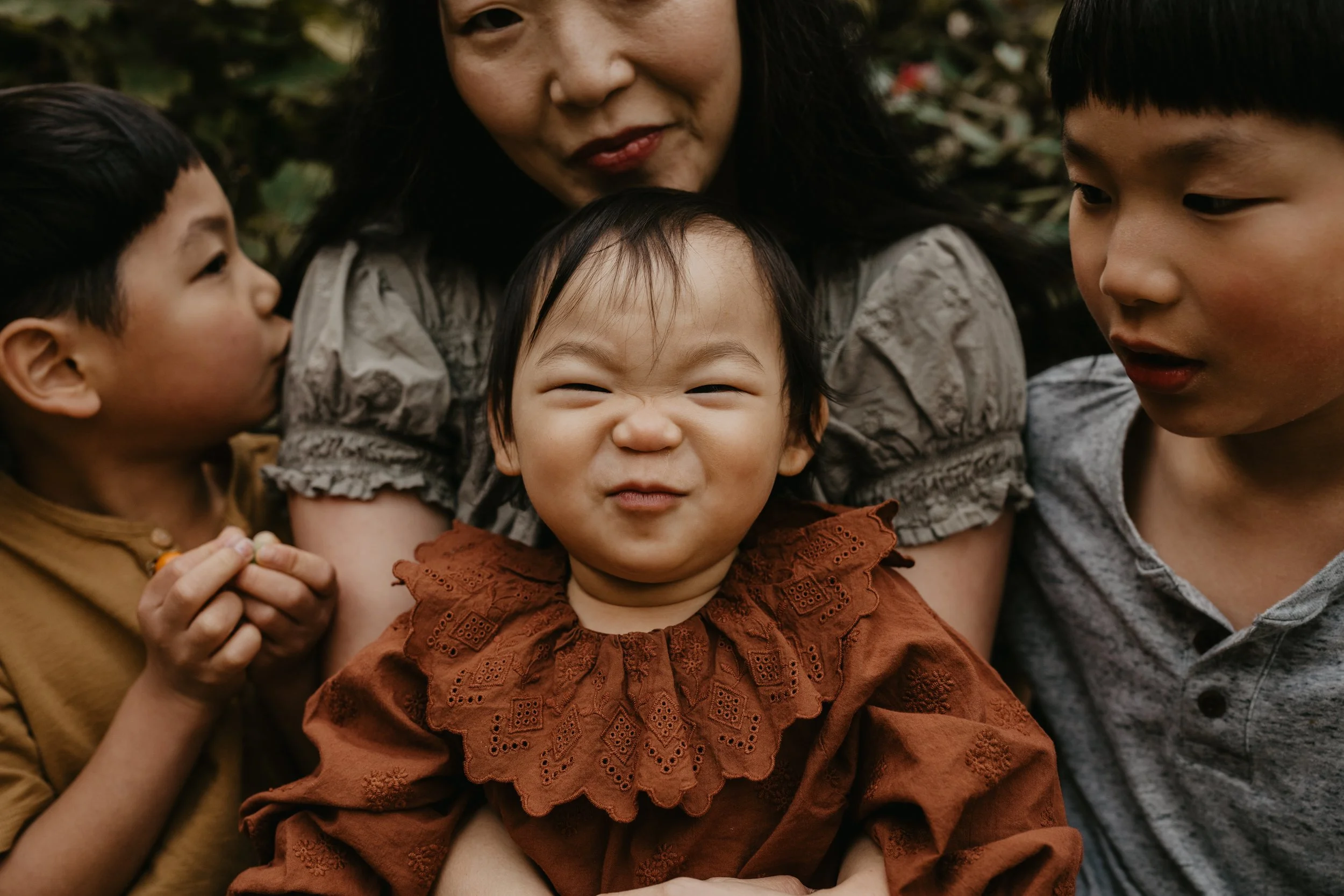 baby making a funny face at the camera while older siblings kiss mom