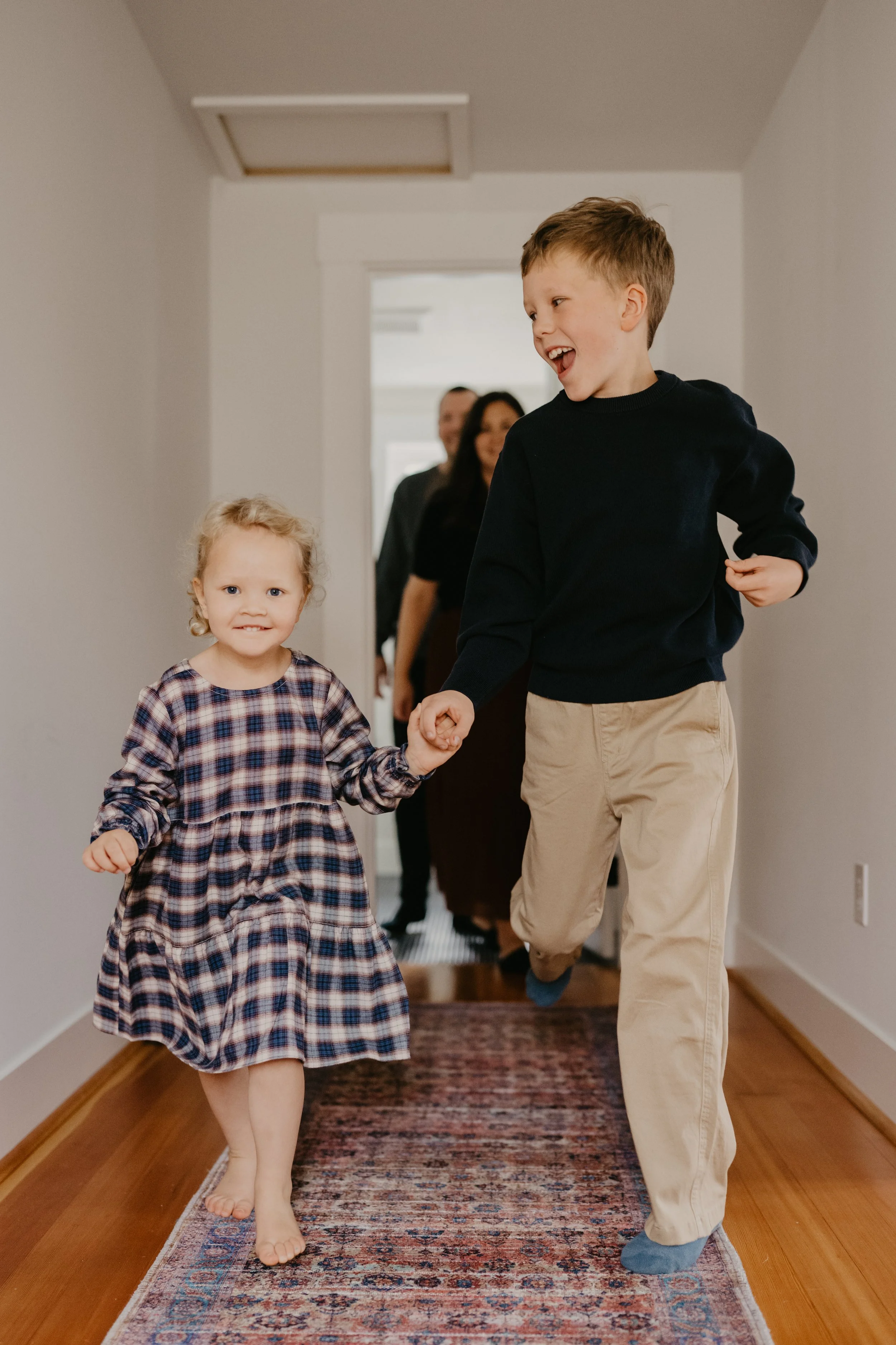 A young boy and girl walking hand-in-hand down a hallway, smiling. The boy is dressed in a black sweater and khaki pants, while the girl is wearing a plaid dress. In the background, a family is seen following them, smiling.