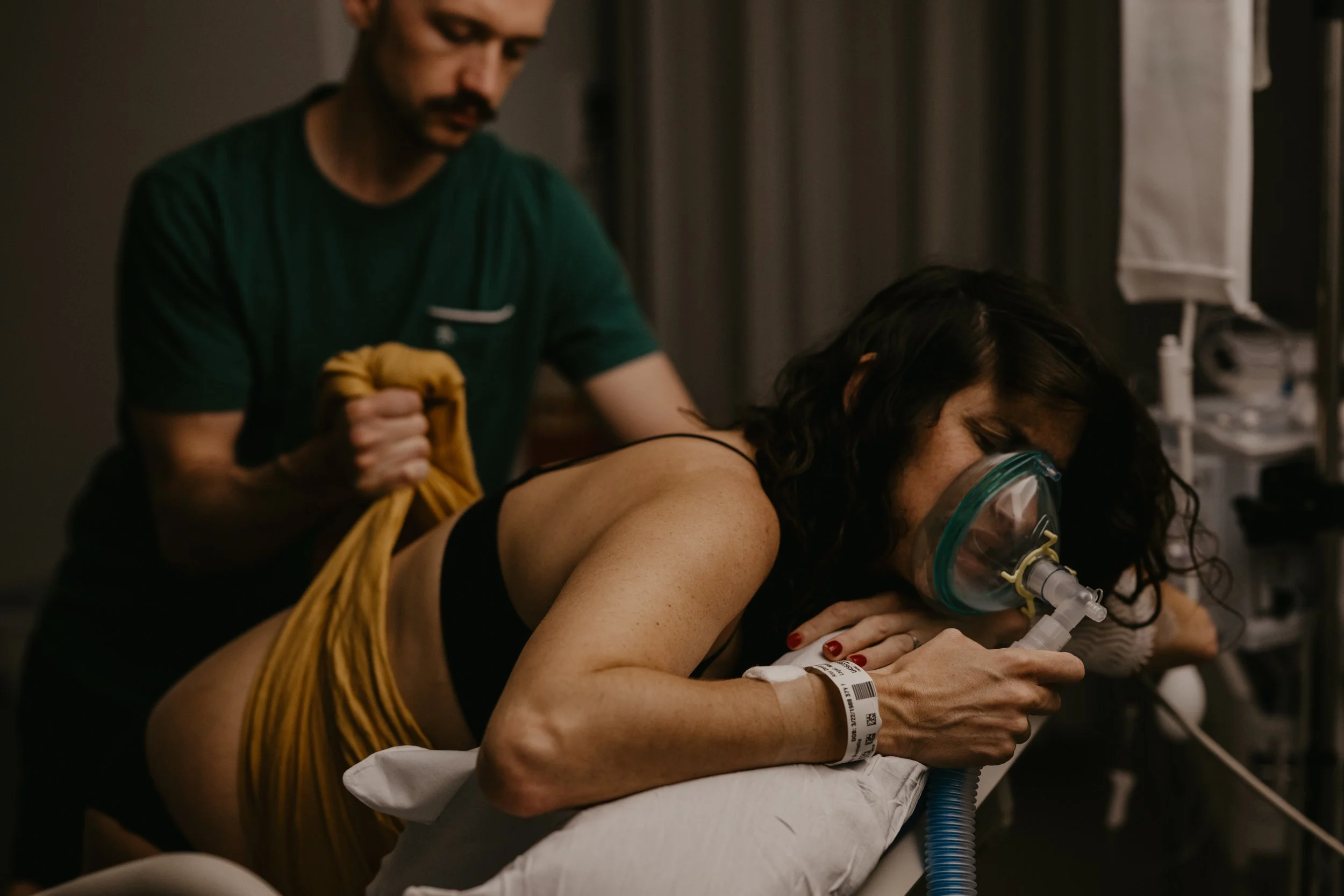 A woman lying on a hospital bed, wearing an oxygen mask, and a green wristband, with a healthcare professional attending to her.