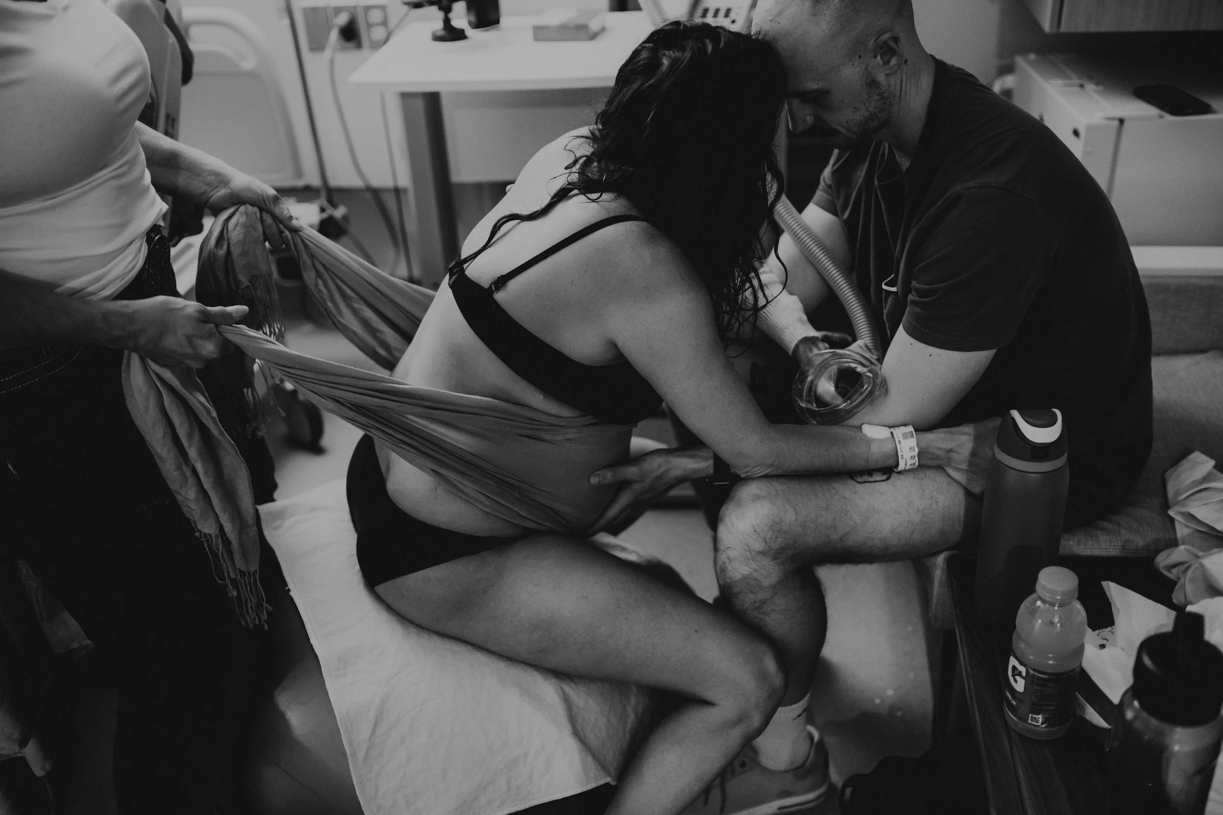 A woman in labor on a hospital bed, wearing black underwear and a hospital wristband, with medical staff assisting her through her childbirth. The scene appears intense and emotional, with the woman leaning forward and a man supporting her.