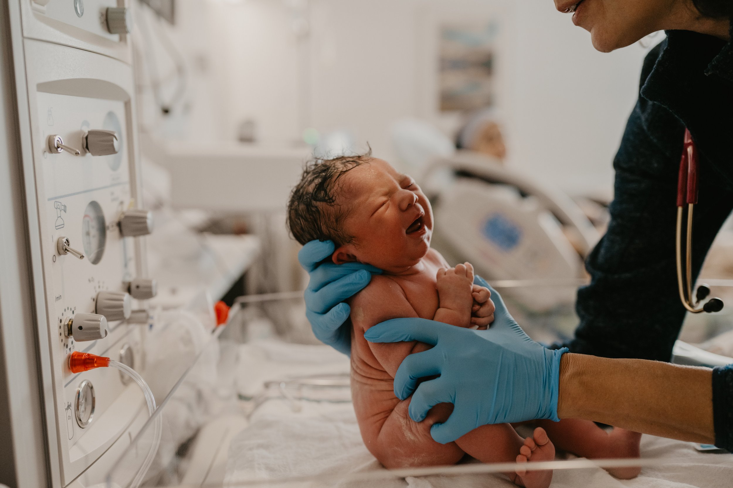 Newborn baby crying during delivery in a hospital, held by a healthcare professional wearing blue gloves, with medical equipment visible nearby.
