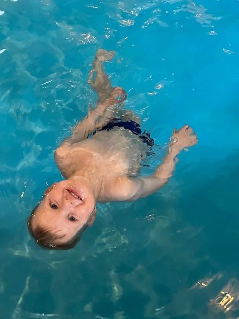 Young child floating and playing in a swimming pool, smiling and looking at the camera.