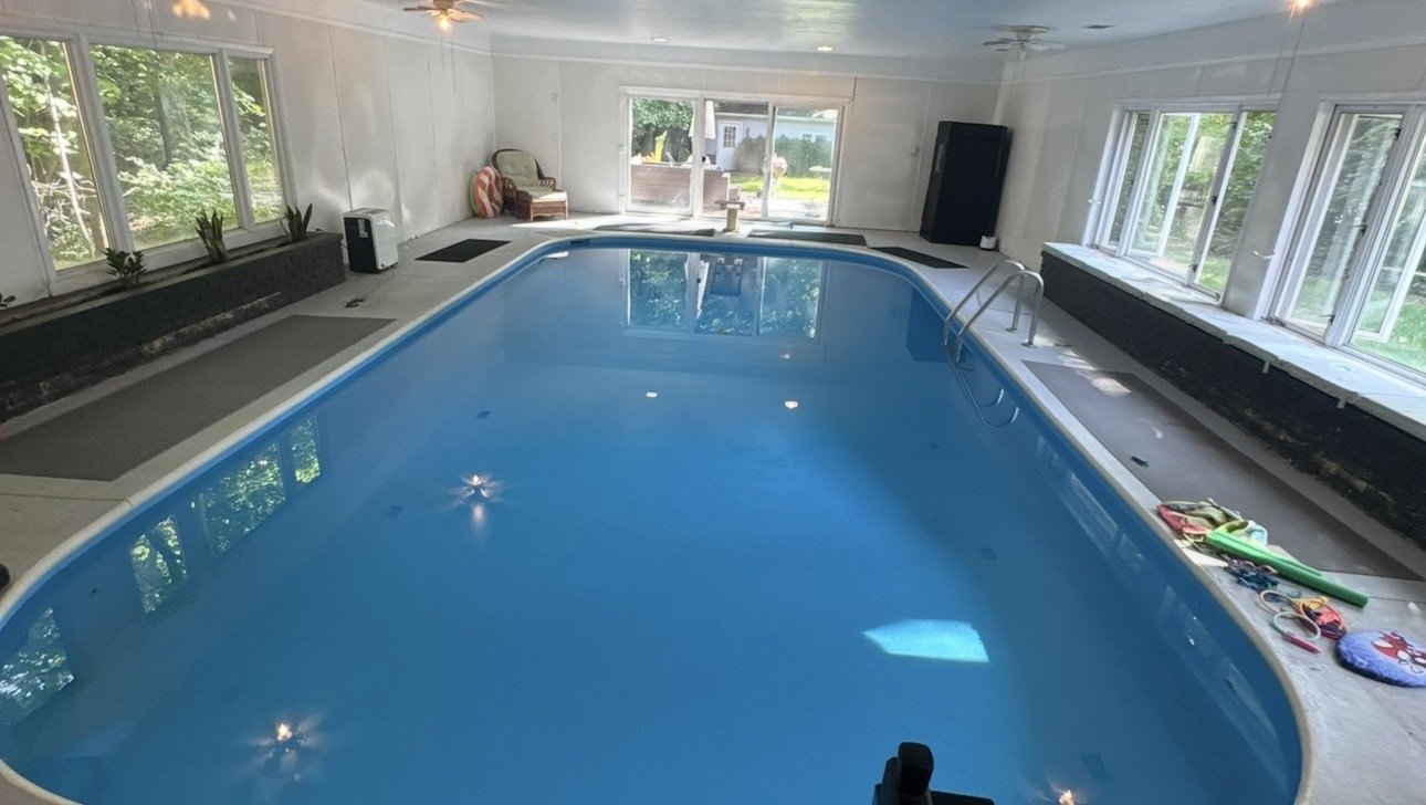 Indoor swimming pool in a room with large windows, with a ladder and surrounded by various pool accessories and furniture.