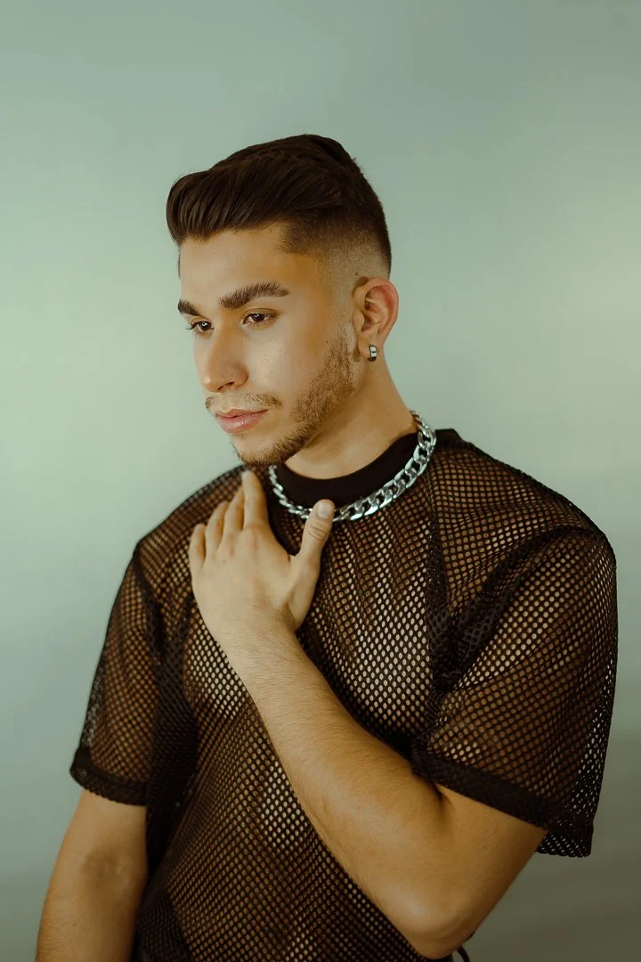 A young man with styled brown hair, wearing a black mesh shirt, a chain necklace, and earrings, holding his hand to his chest against a plain light background.
