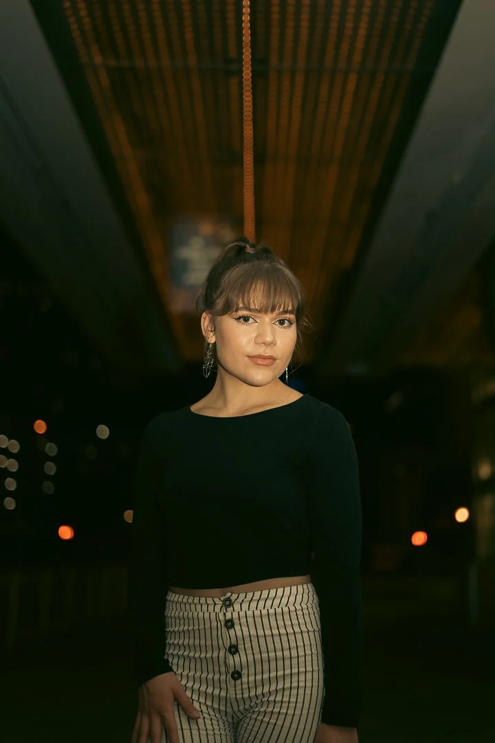 A young woman with brown hair styled in a high ponytail and bangs, wearing a black long-sleeve top and striped high-waisted pants, standing outdoors at night in front of a dark background with blurred lights.
