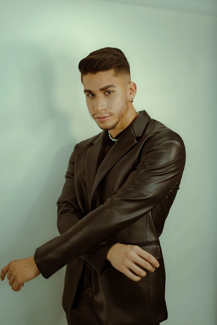 A young man with styled hair and earrings, wearing a black leather jacket, posing against a plain light green background.