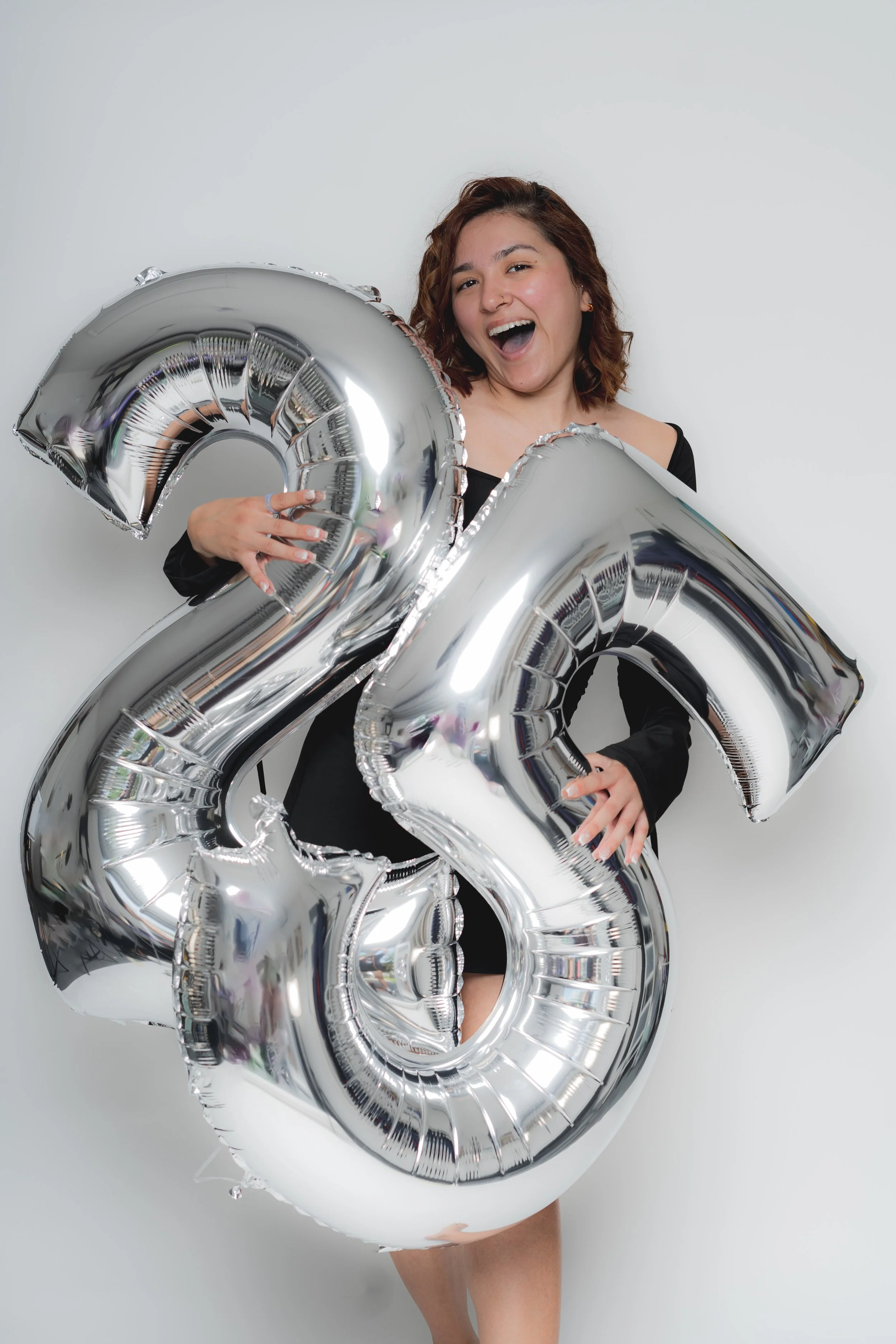 A woman holding large silver balloons shaped as the numbers 2 and 3, celebrating a 23rd birthday or anniversary, standing against a plain white background, smiling and looking joyful.