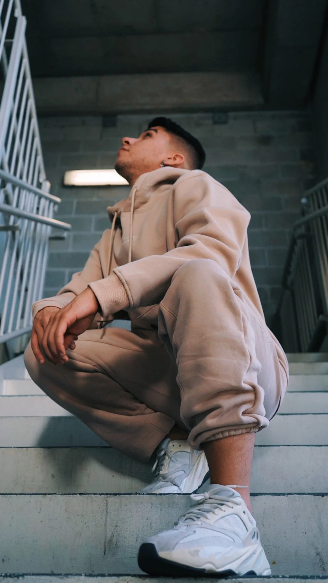A young man crouching on concrete stairs indoors, wearing a beige hoodie, matching sweatpants, and white sneakers, looking up.