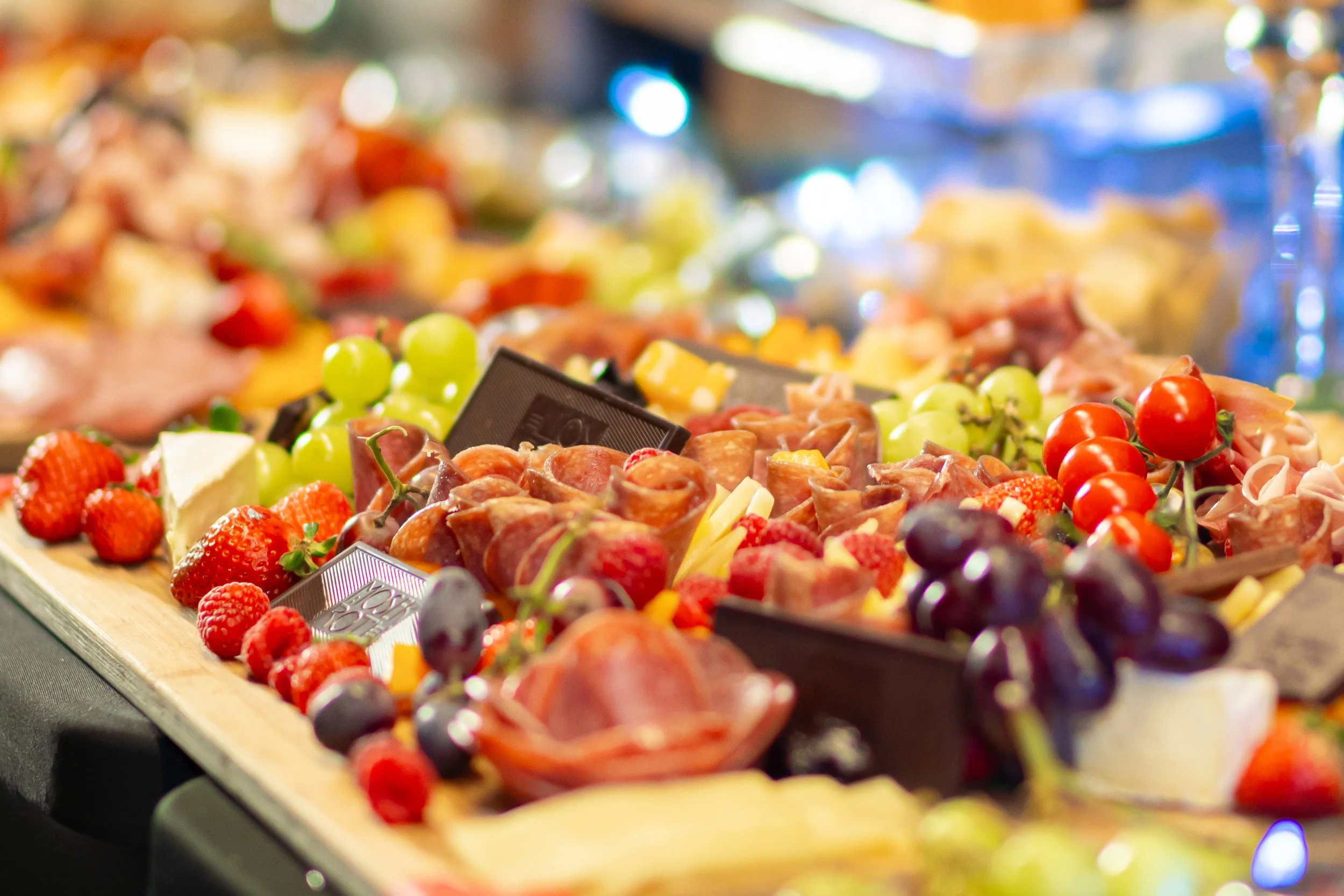 Assorted appetizers on a platter including strawberries, grapes, cherry tomatoes, cheese, and slices of deli meats at a buffet or party setting.