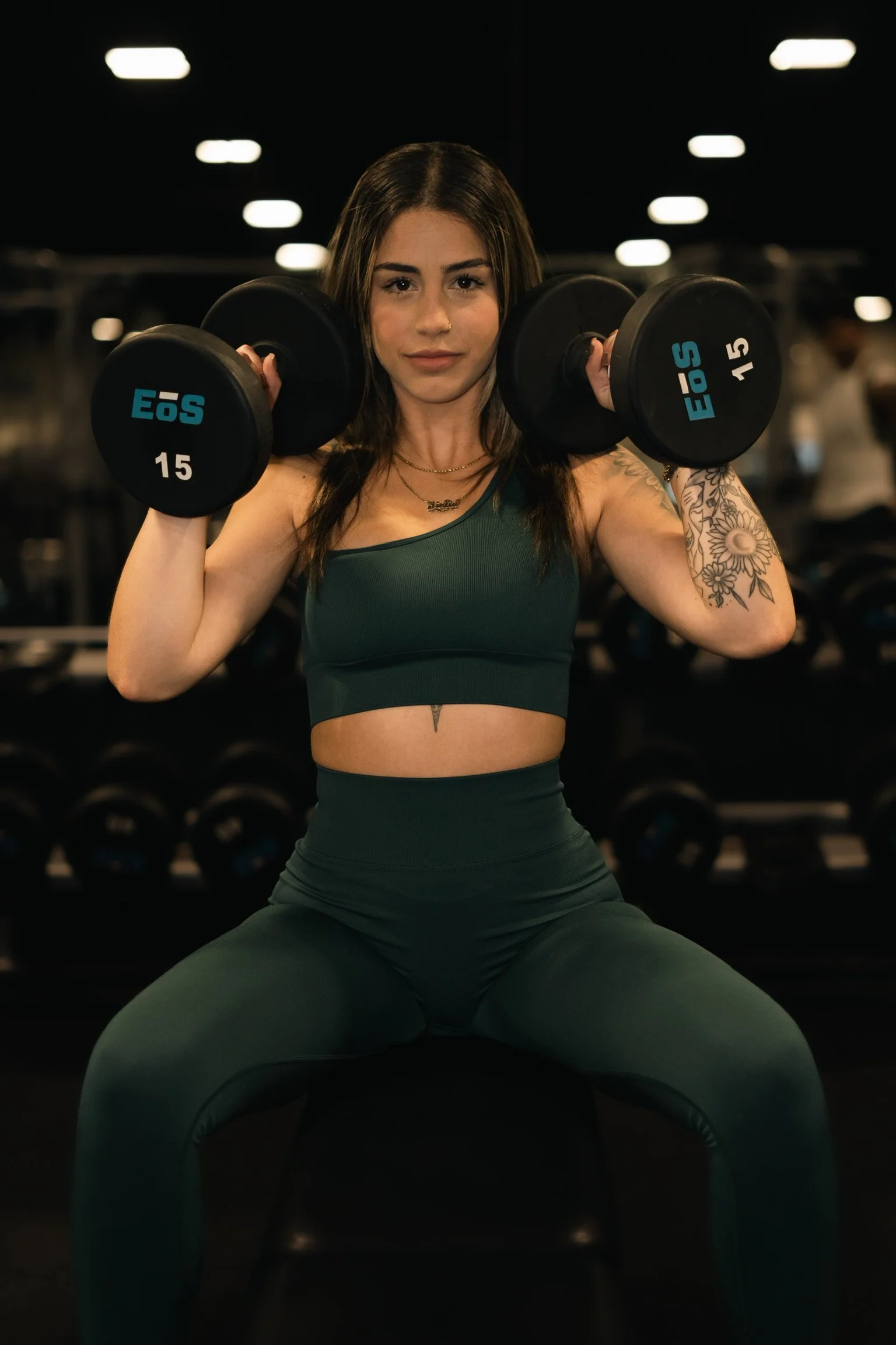 A woman lifting dumbbells in a gym, wearing a dark green sports bra and leggings, with tattoos on her left arm and a tattoo on her torso, focused on her workout.