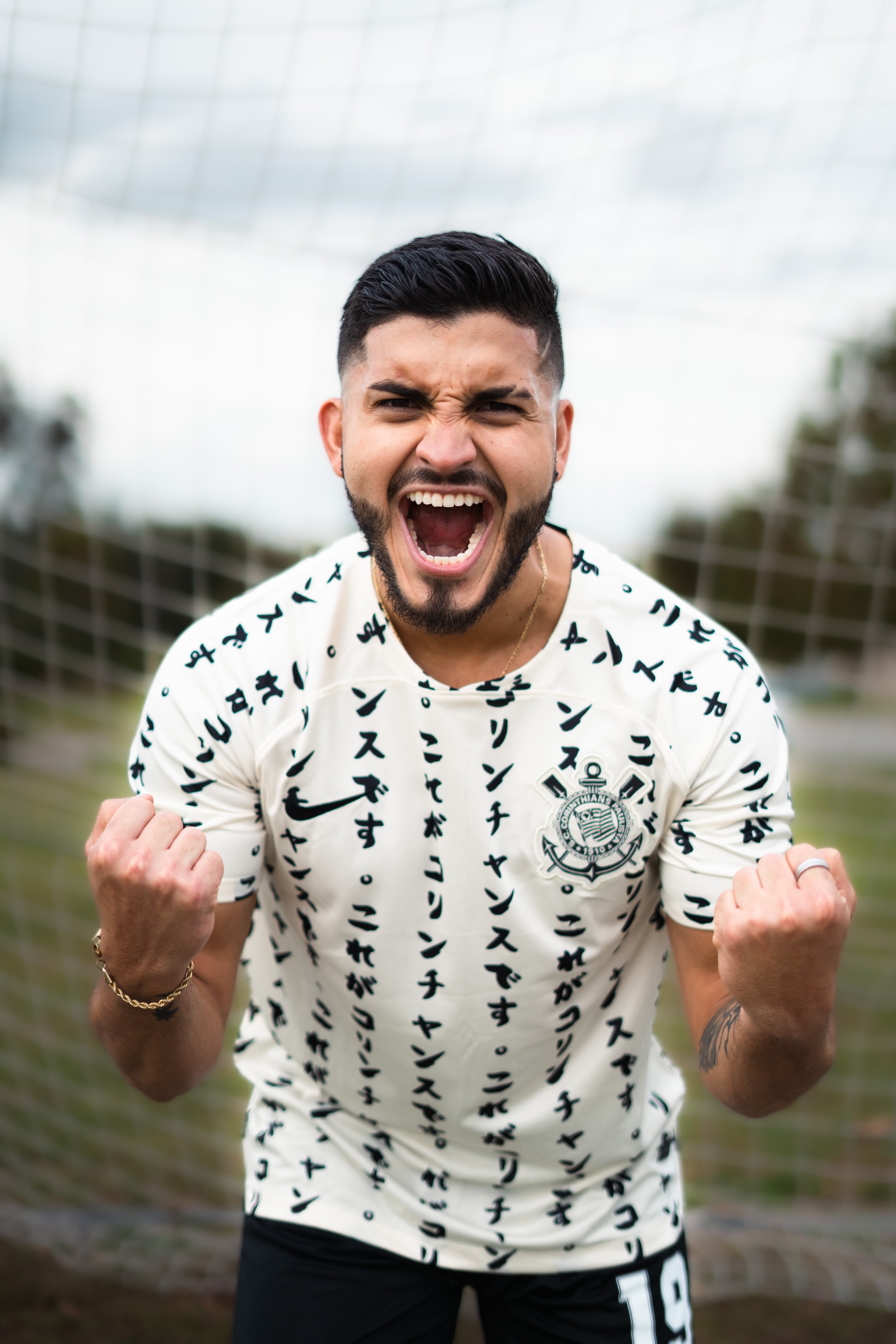 A man with short dark hair and a beard, wearing a white shirt with black Japanese characters and a crest, is celebrating with a fist pump and shouting excitedly on a soccer field with a net in the background.