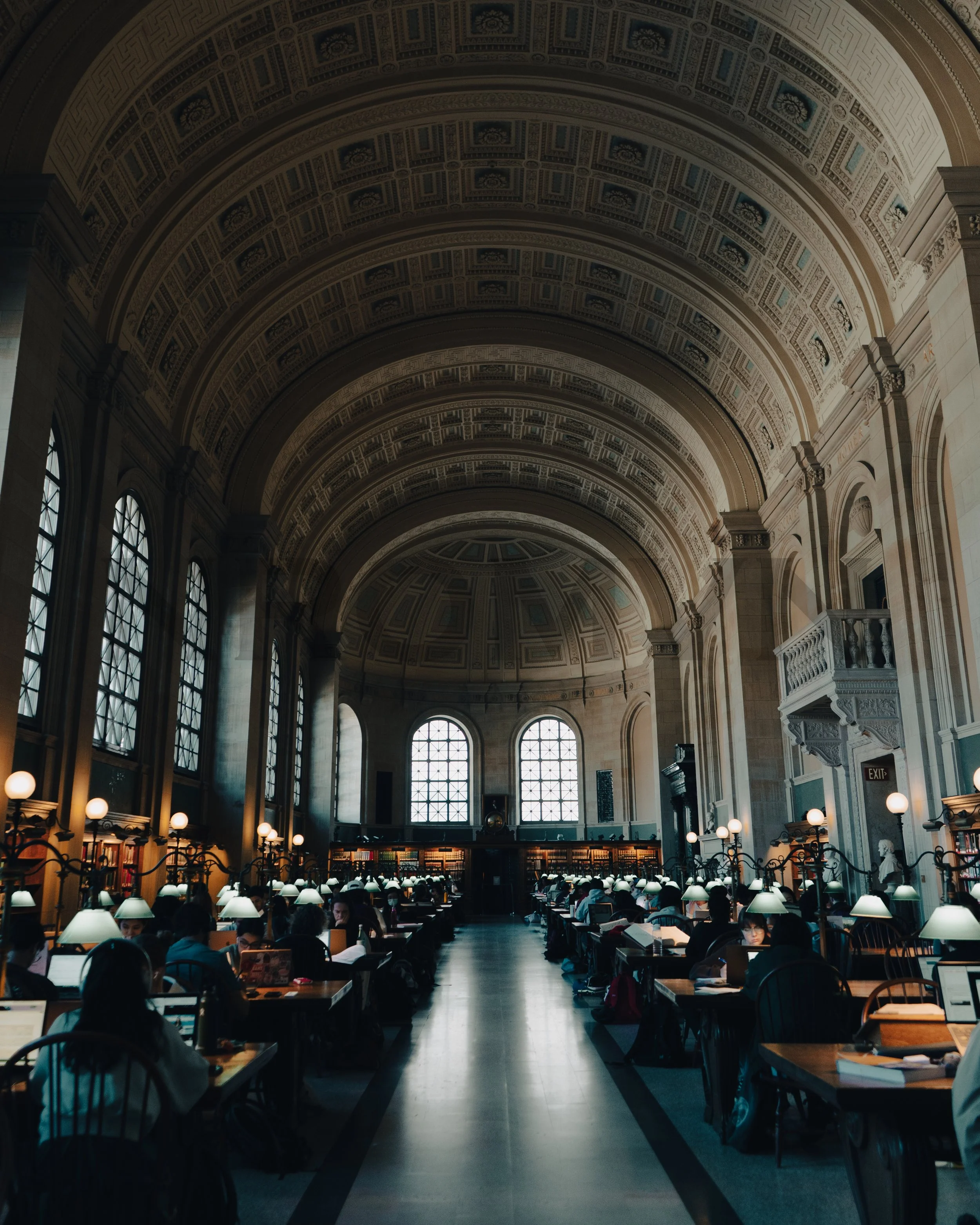 A grand library interior with high arched ceilings, large windows, and rows of tables with people studying or working.
