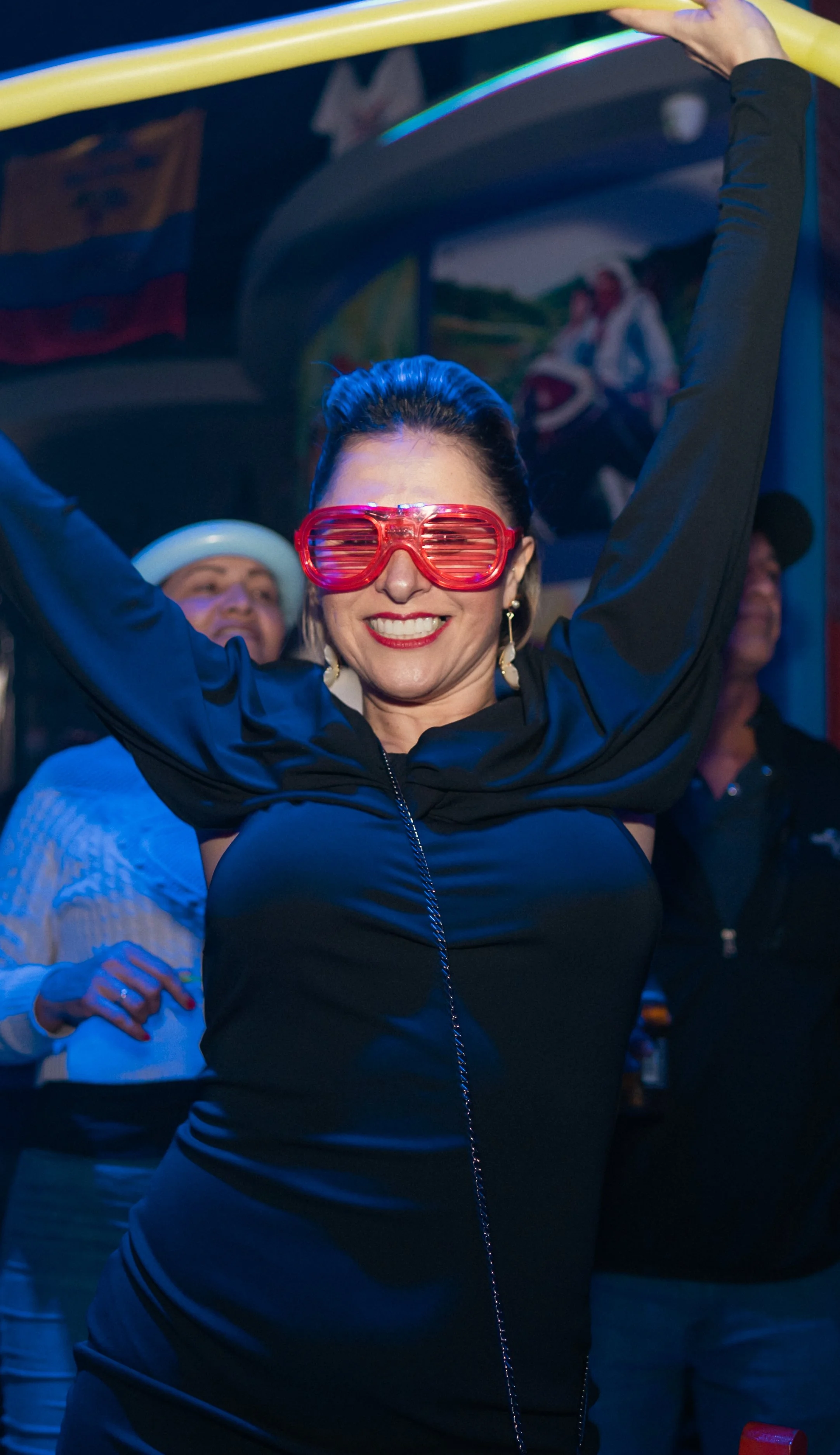 A woman dancing with a wide smile, wearing red futuristic glasses, a black dress, and earrings, with others in the background in a festive environment.