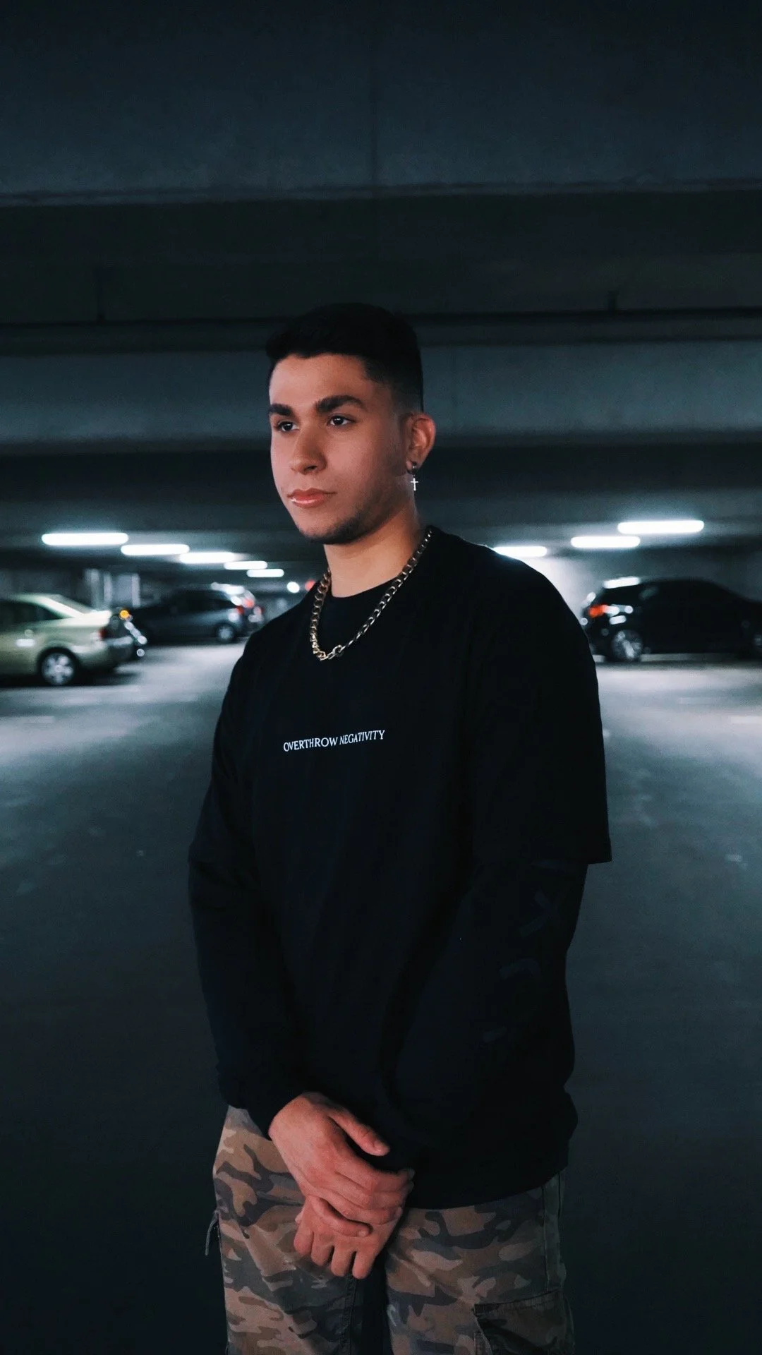 Young man standing in a parking garage at night, wearing a black shirt with white text, camouflage pants, and jewelry.