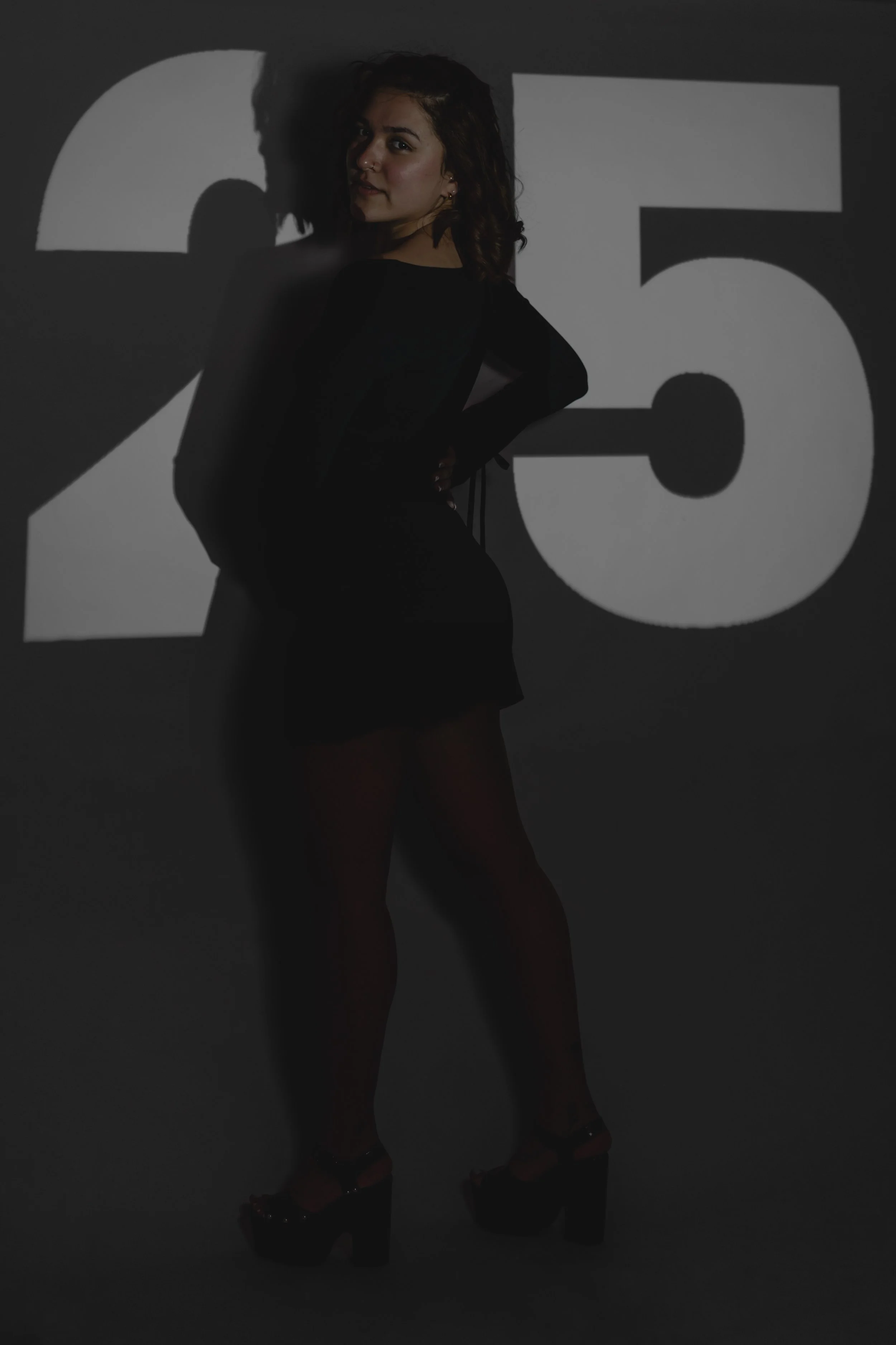 A woman standing in front of a wall with the number 25 projected onto it, wearing a black dress and high heels.