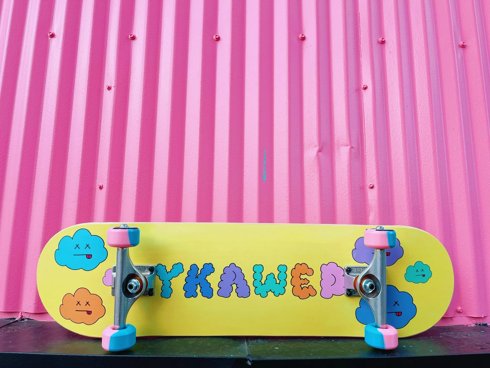 A yellow skateboard with colorful clouds and the word "YKAWE" on it, set against a pink corrugated metal wall.