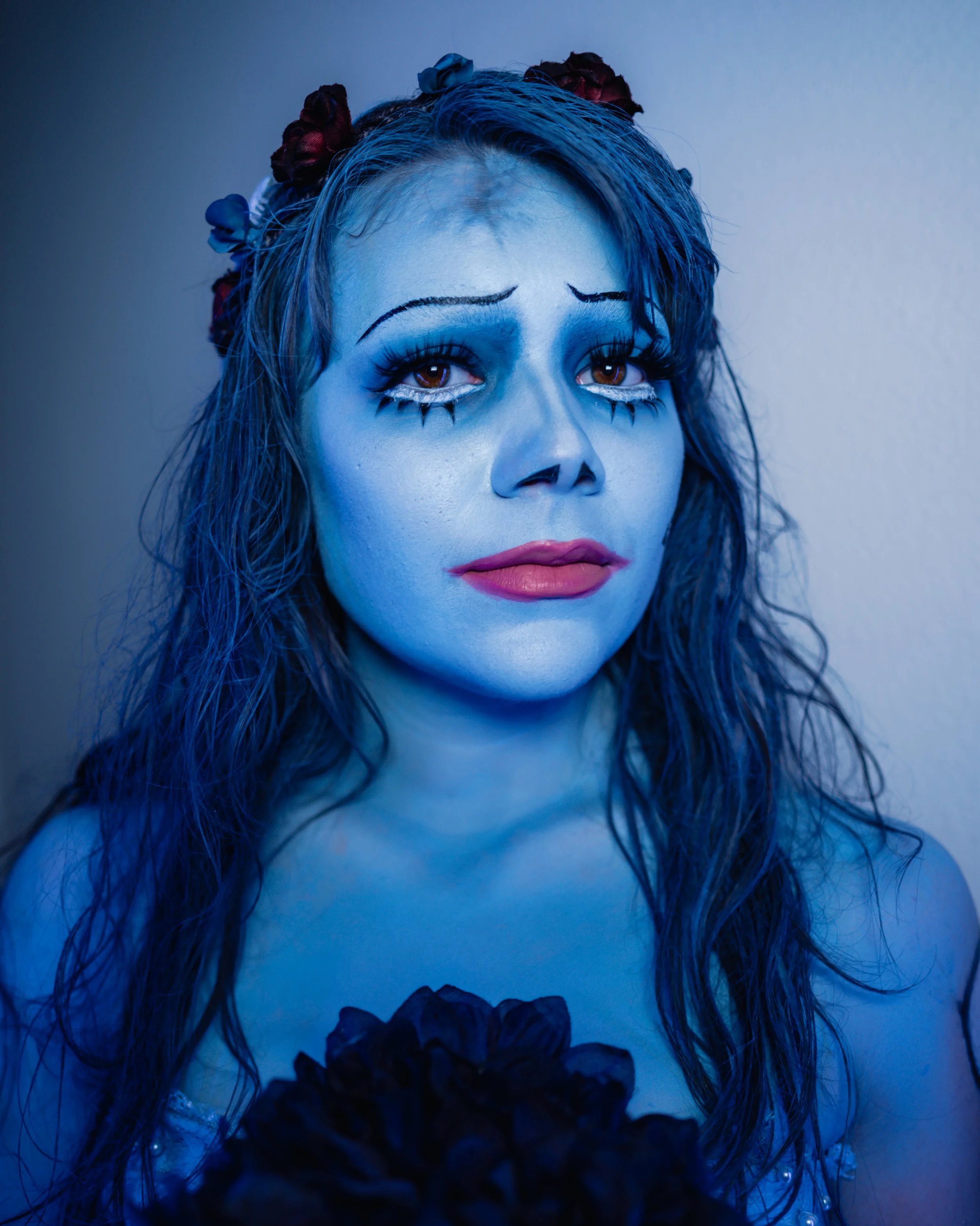 A woman dressed as a sad clown with blue makeup, dark curly hair with flowers, and black makeup accents around her eyes and lips.