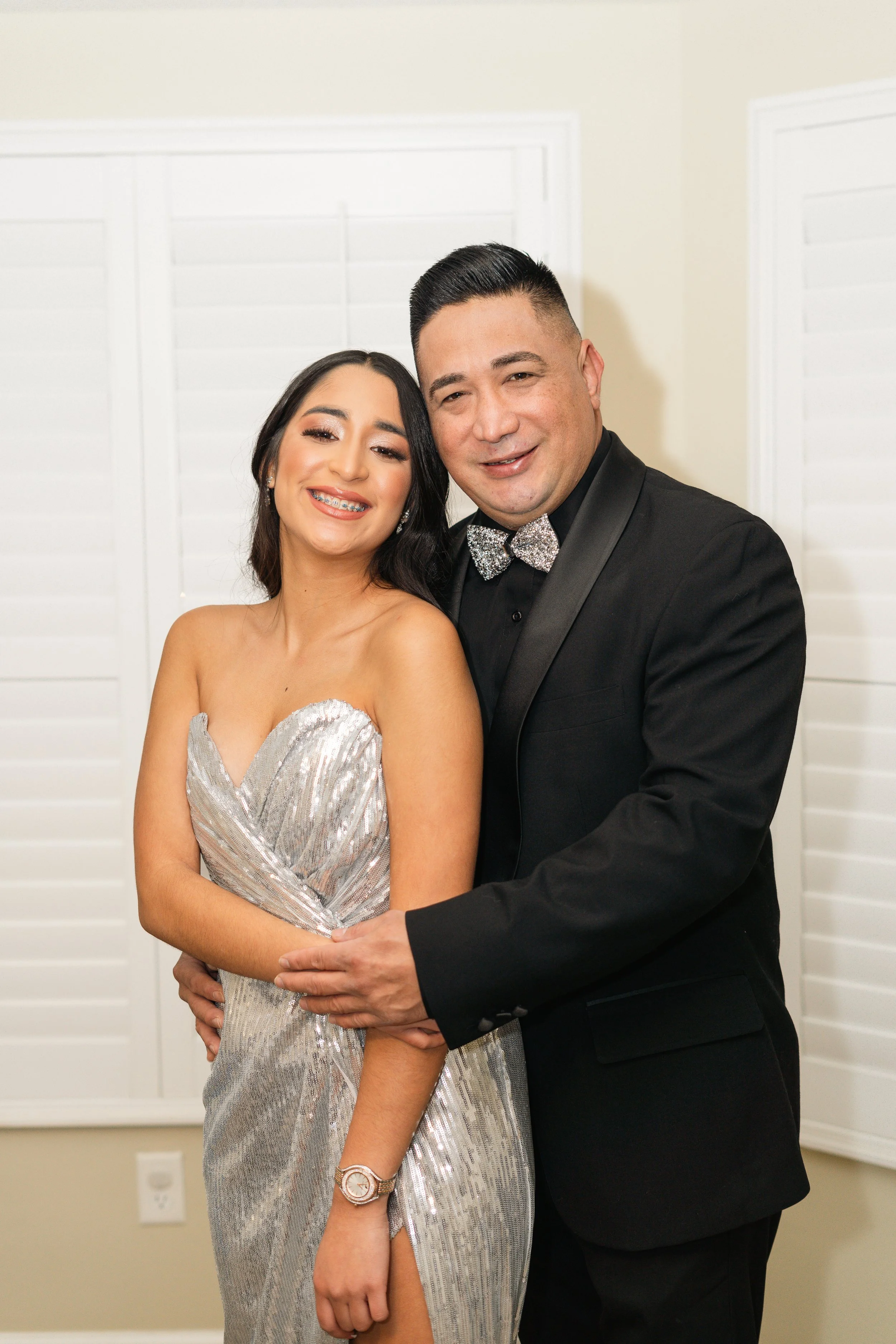 A happy couple dressed in formal attire, the woman in a silver strapless gown and the man in a black tuxedo with a sparkly bow tie, standing close together and smiling in front of white window shutters.