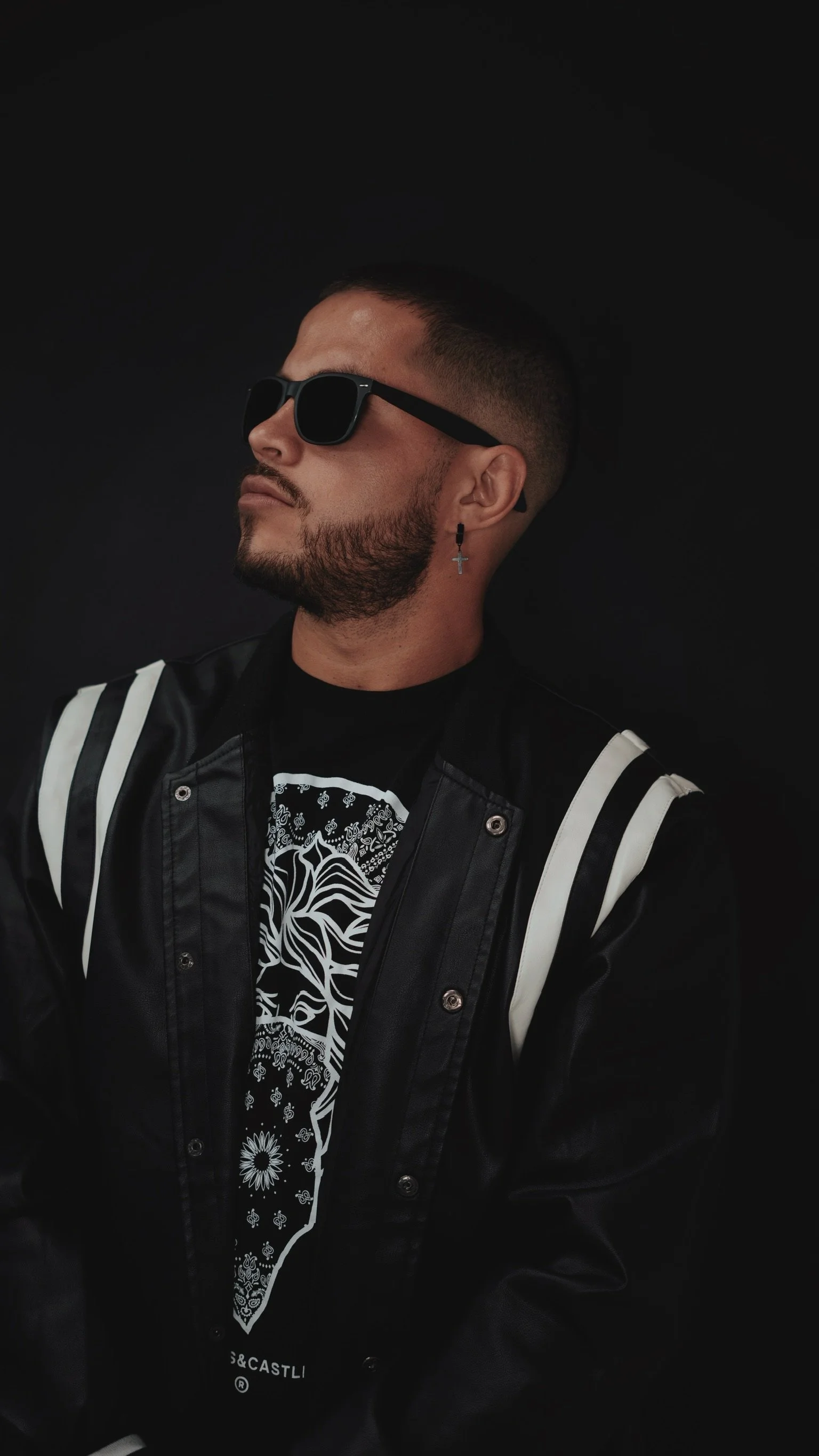 A young man with a beard and short hair, wearing black sunglasses, a black jacket with white stripes on the shoulders, and a black T-shirt with white graphic design, posing against a black background.