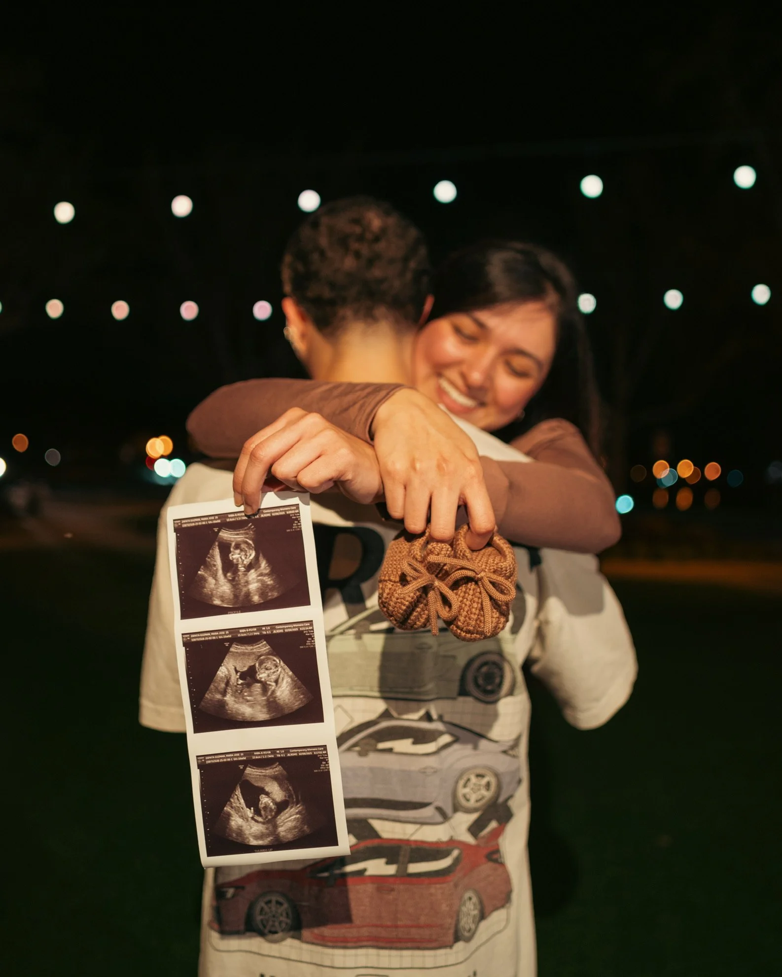 A woman and a man are hugging. The woman is smiling with eyes closed and holding an ultrasound image. The background shows blurred lights at night.