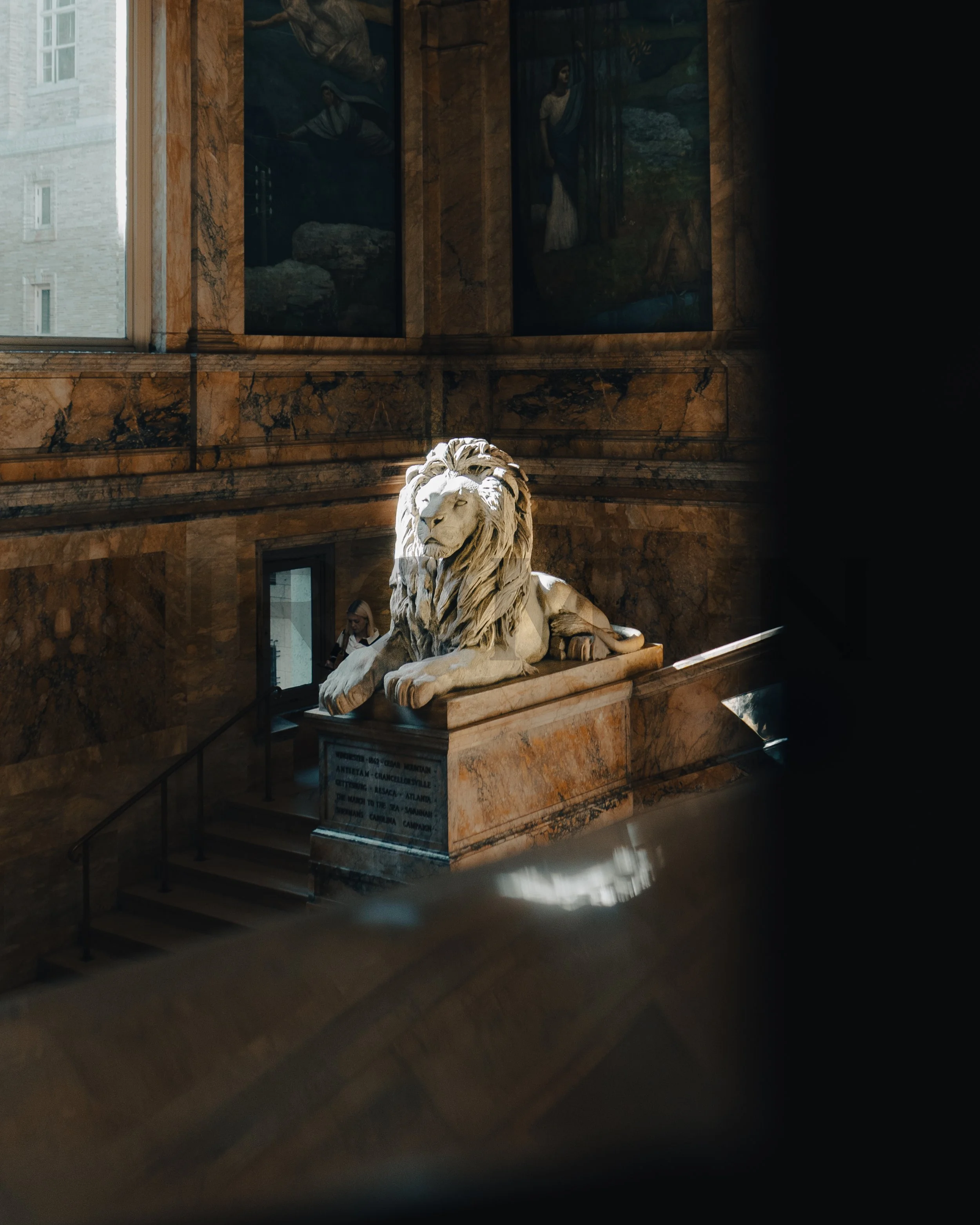 Marble lion statue inside a grand building with marble walls, illuminated by sunlight, with paintings on the wall behind it.