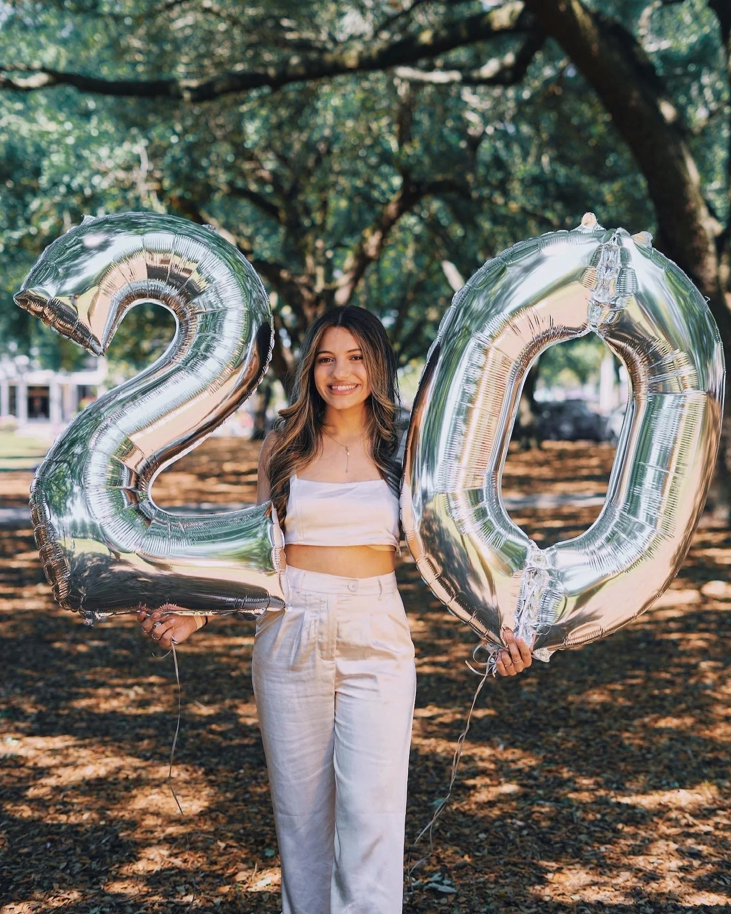 A woman holding large silver balloons shaped as the numbers '2' and '0' in an outdoor park with trees in the background, celebrating a 20th birthday or anniversary.