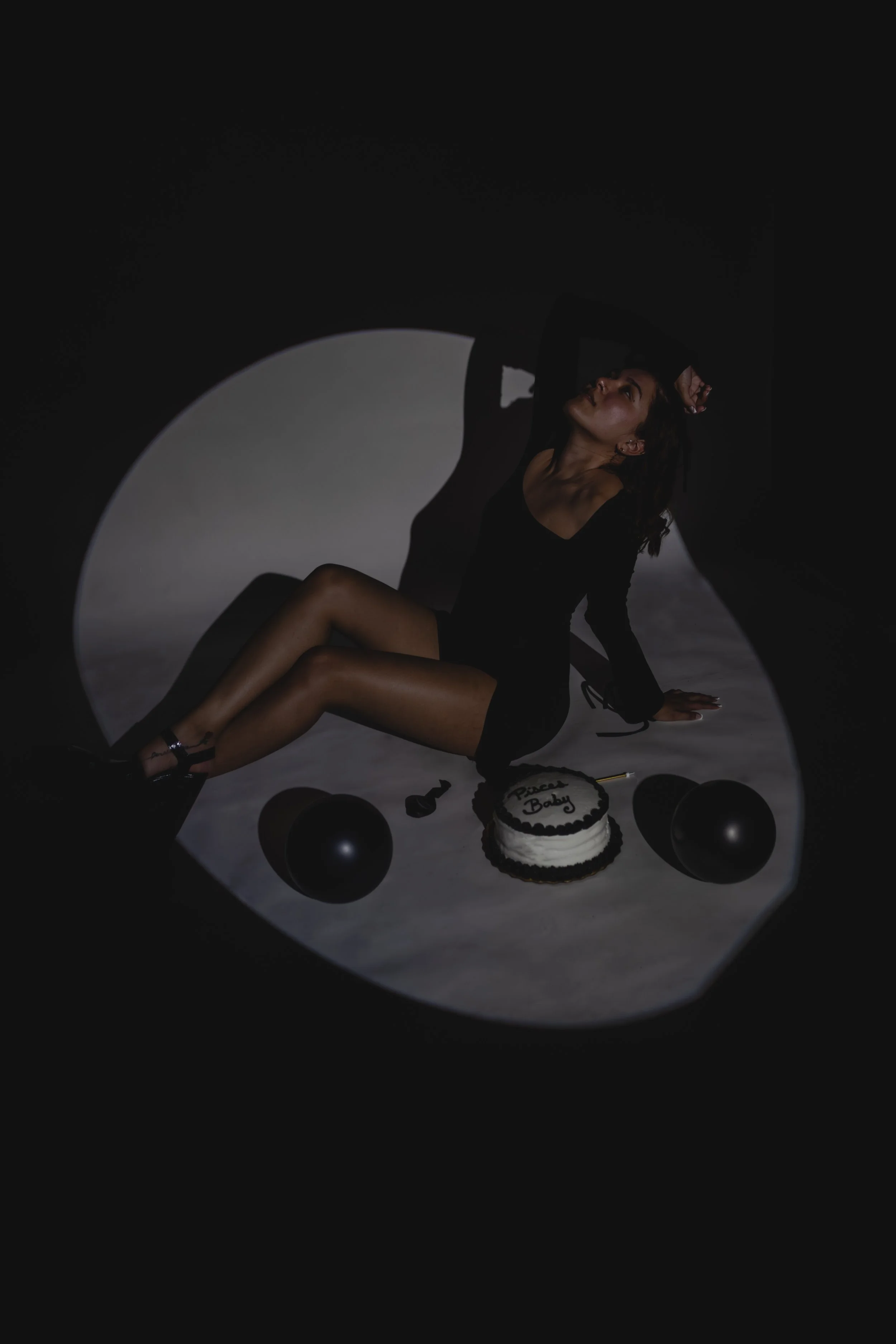 A woman in black sitting on the floor with a birthday cake, balloons, and party items, in low lighting with a spotlight creating a shadow circle.