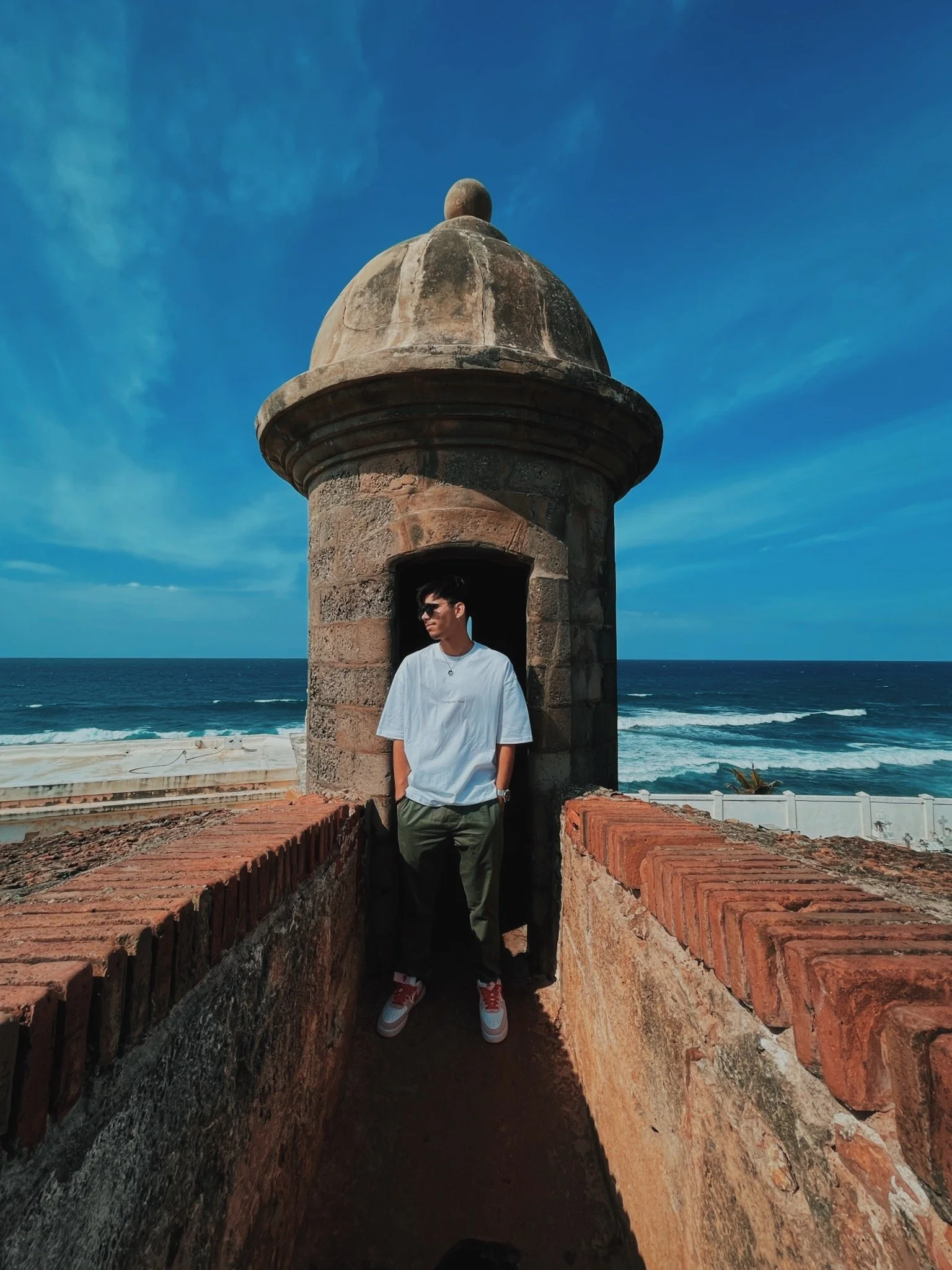 A young man wearing sunglasses, a white t-shirt, and green pants standing inside a small stone tower by the ocean, with a blue sky and ocean waves in the background.