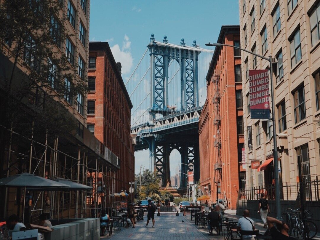 View of Manhattan Bridge seen from a busy city street in Brooklyn, New York City, with buildings on either side, people sitting and walking, and outdoor seating areas.