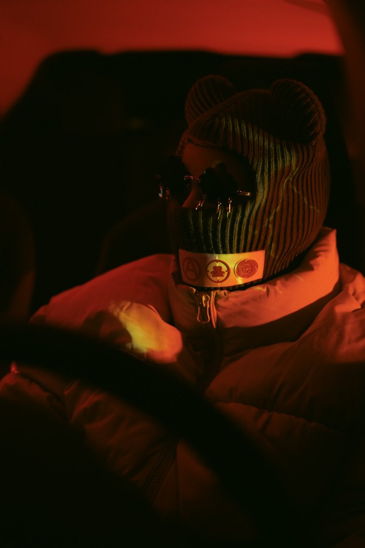 A person wearing a striped knit hat, sunglasses, and a white puffy jacket, sitting in a dimly lit environment with red lighting.