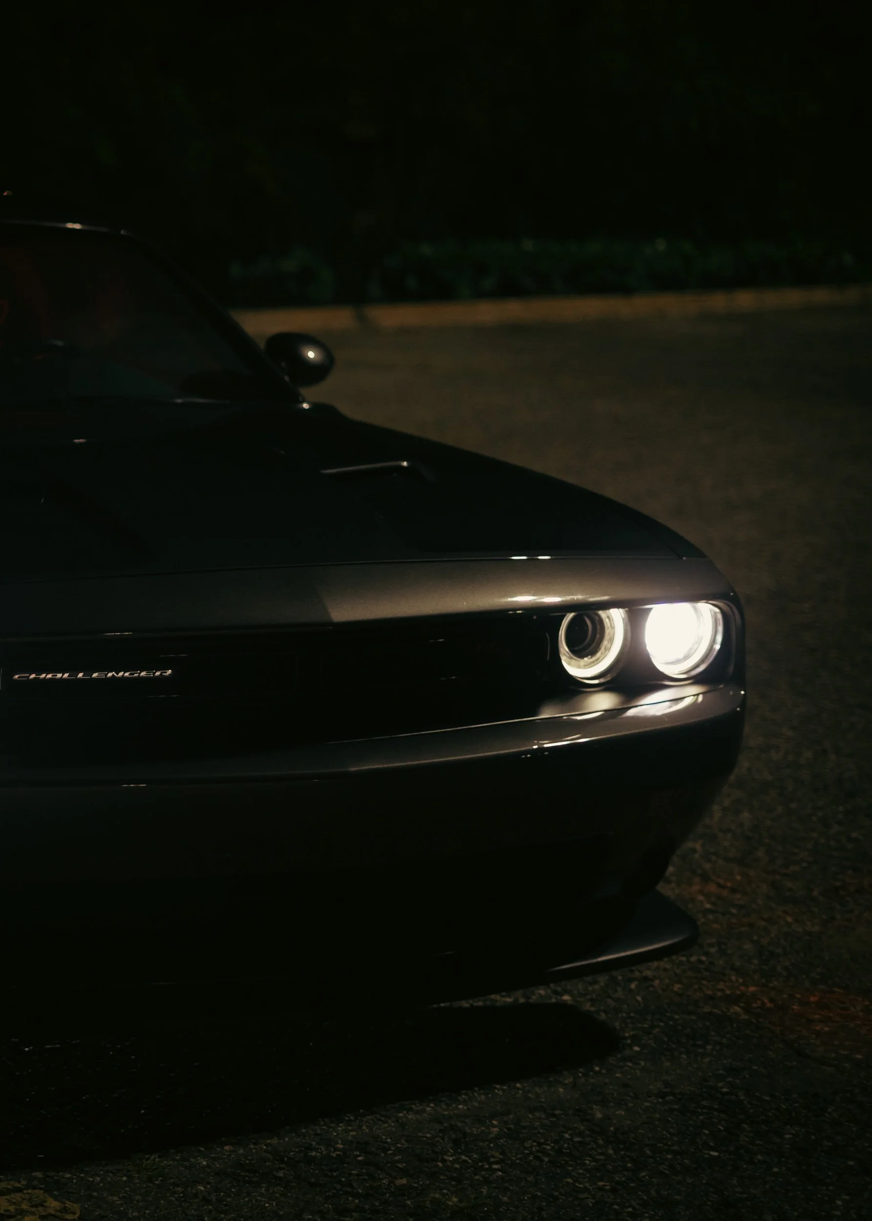 Close-up of the front of a black Dodge Challenger with illuminated headlights, parked on a dark street at night.