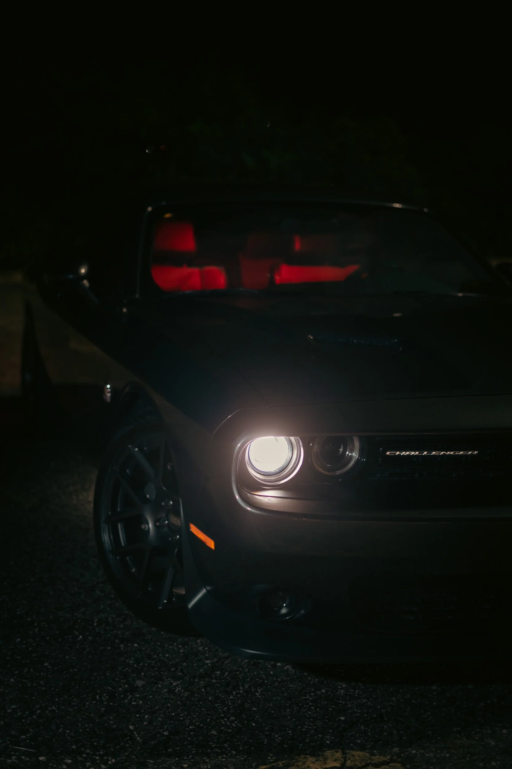 Black Dodge Challenger car at night with red interior lights and one headlight on, parked outdoors on a dark road.