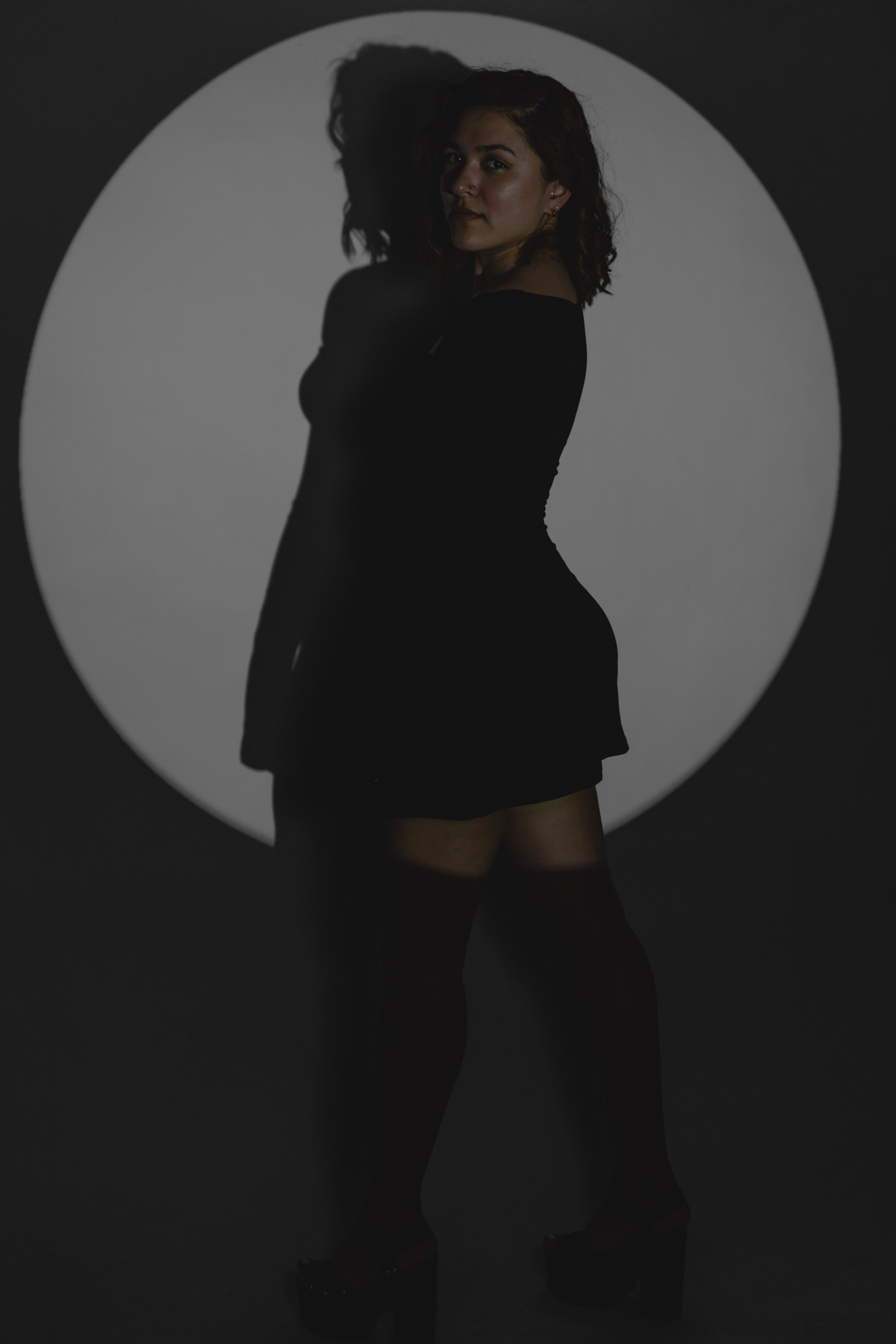 A woman standing in front of a spotlight with a circular shadow behind her. She is wearing a black dress, patterned stockings, and high-heeled shoes, looking at the camera with a confident expression.