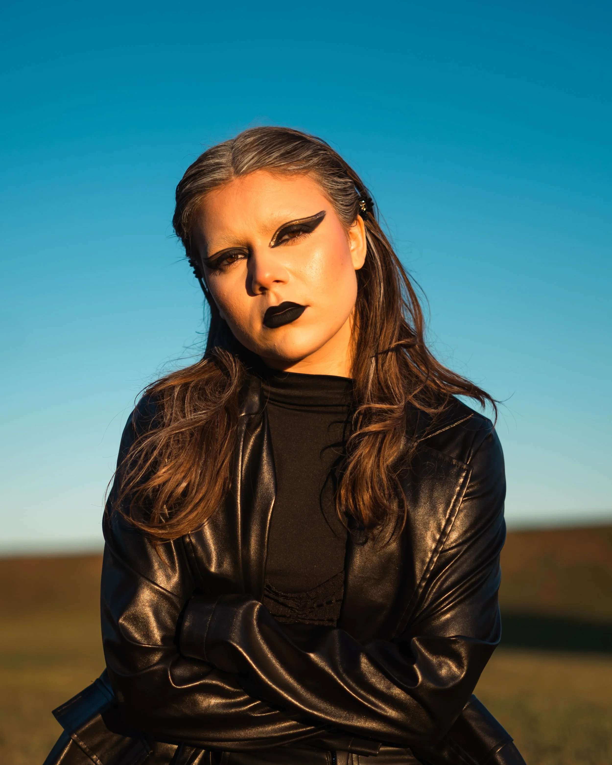 A woman with dramatic black makeup, including bold eyeliner and black lipstick, wears a black leather jacket and a black top, standing outdoors against a clear blue sky.