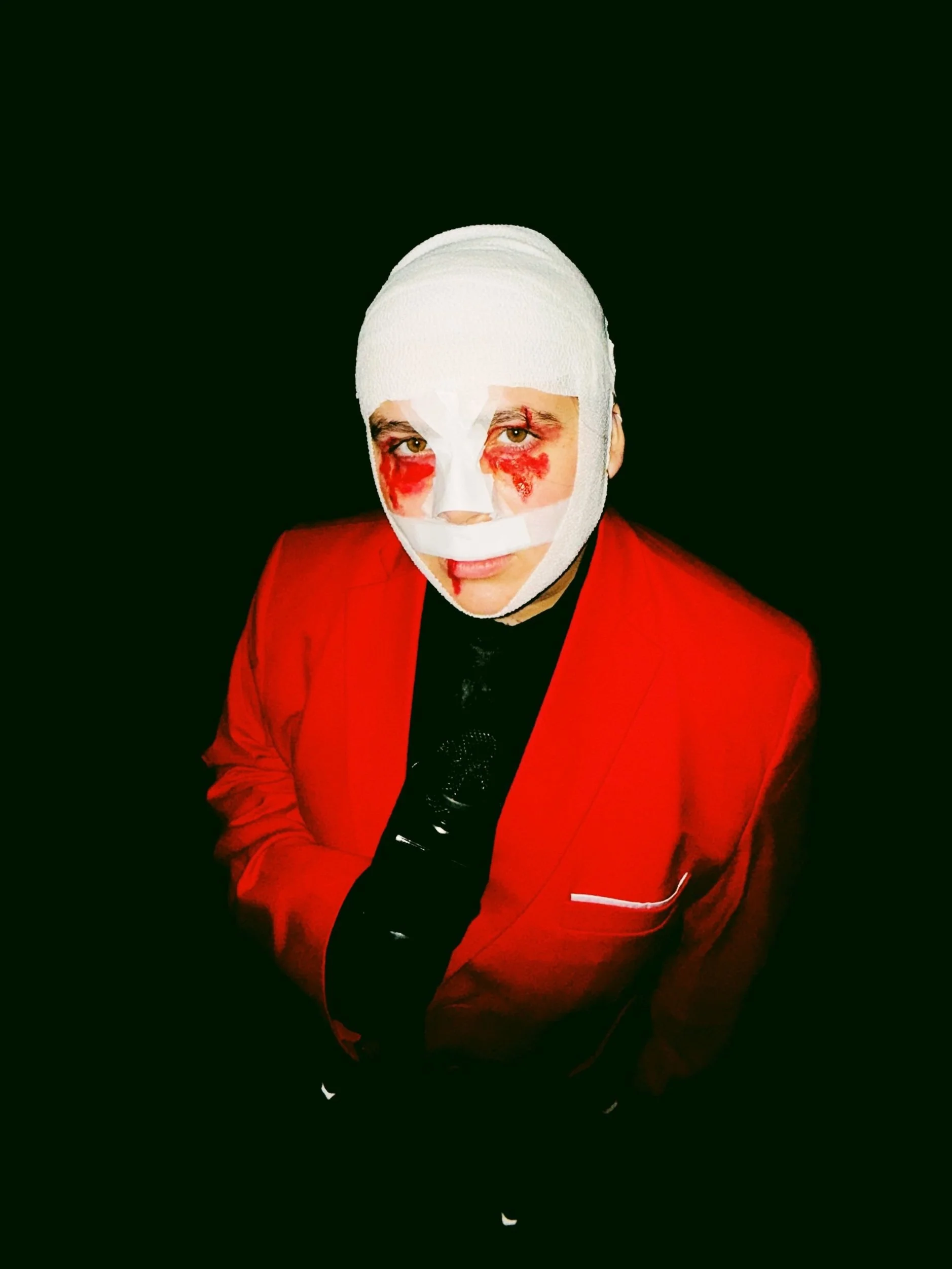 Person with white bandages around head and face, red eyeshadow, blood streaks on face, wearing a red blazer and black clothing, against a black background.