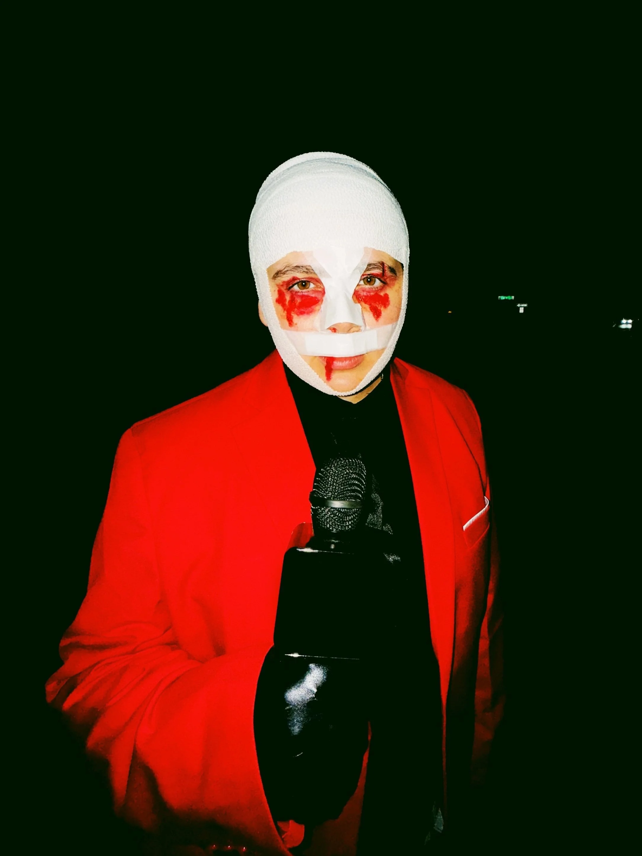 Person with face painted and bandaged head, wearing a red jacket, holding a microphone, standing in a dark background.